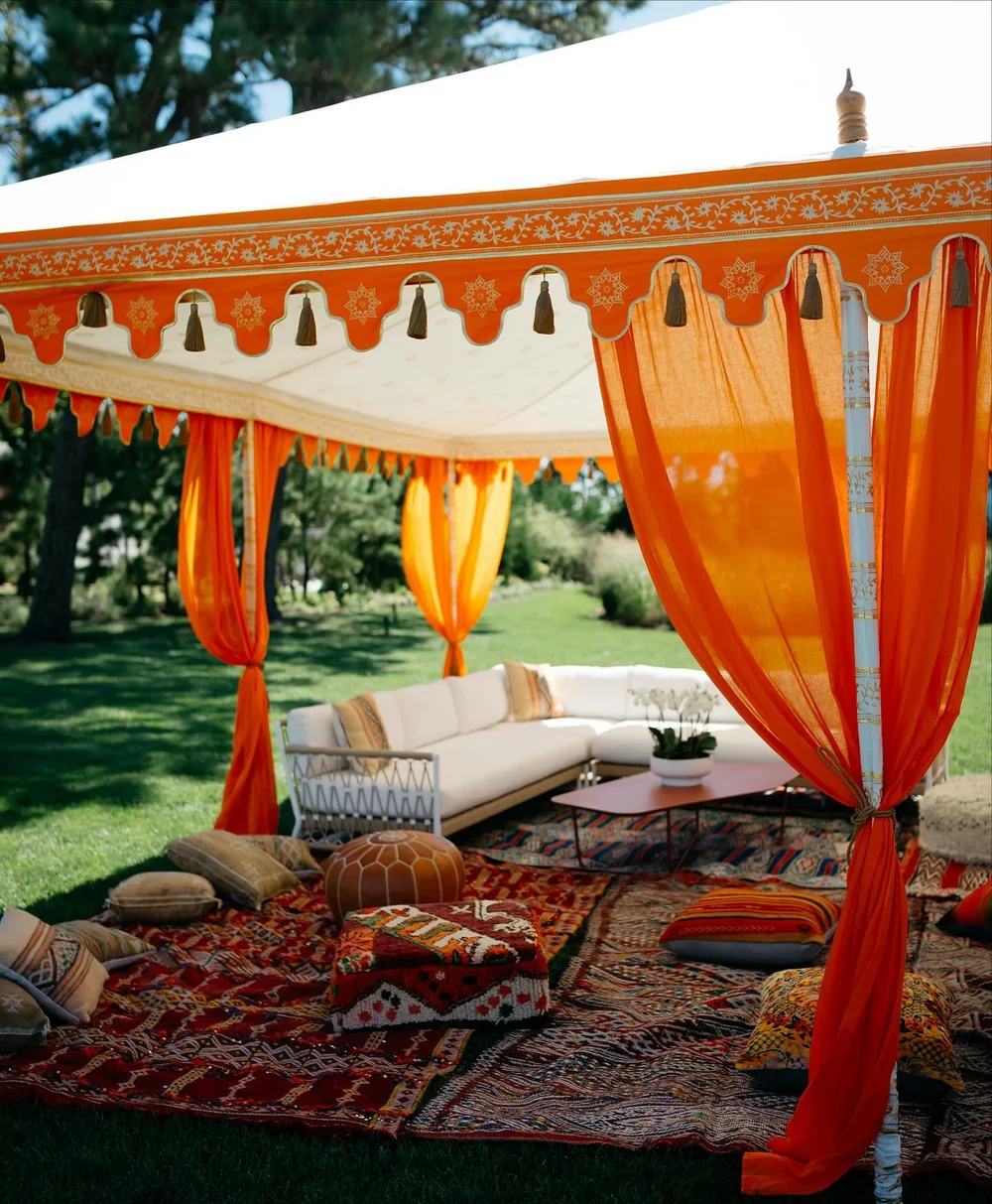 White Moroccan tent with orange drapes, patterned rugs and low lounge seating outdoors