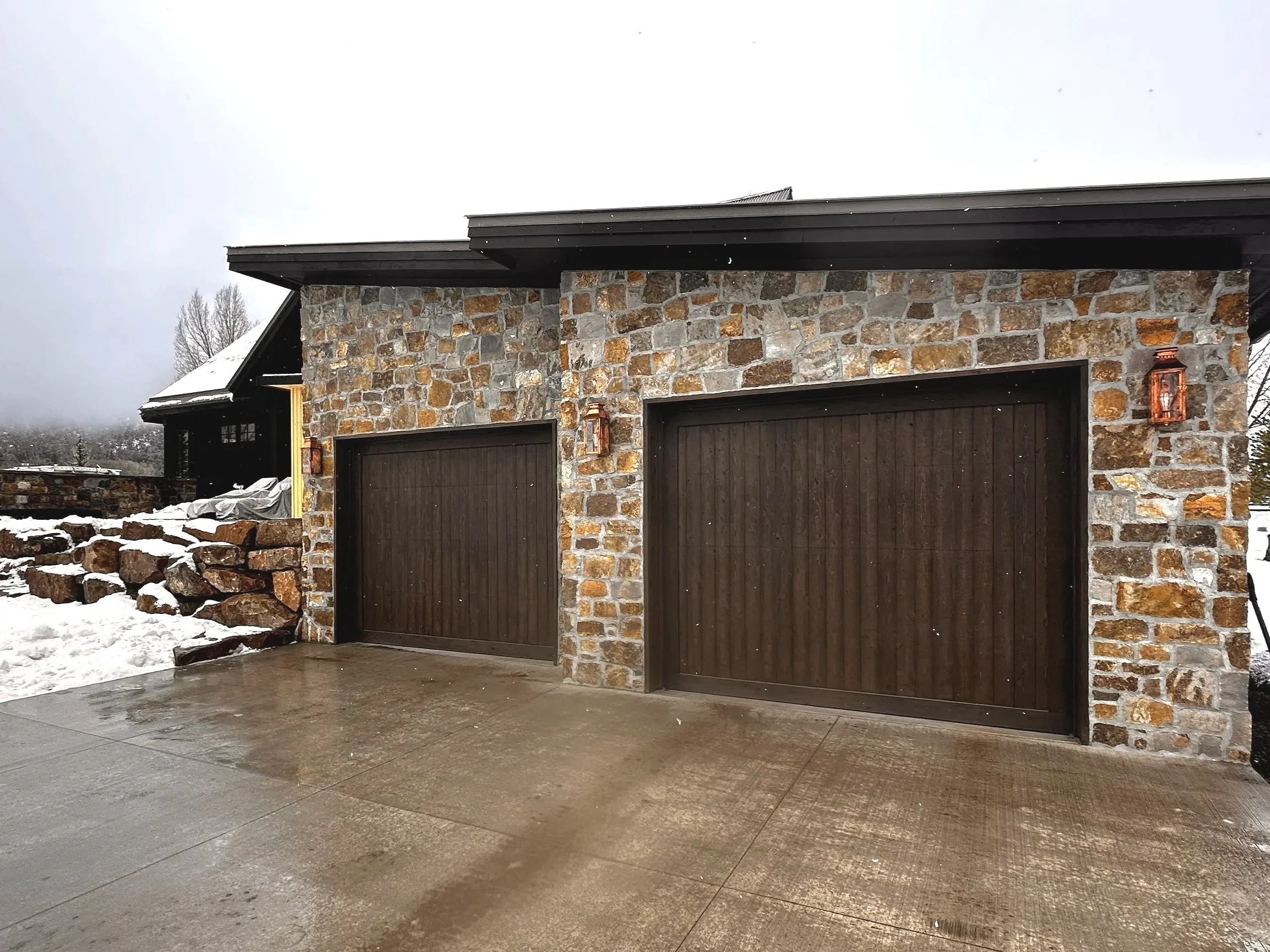 Stone garage with two wooden doors, outdoor wall-mounted lanterns, wet concrete driveway, snowy surroundings, cloudy sky.