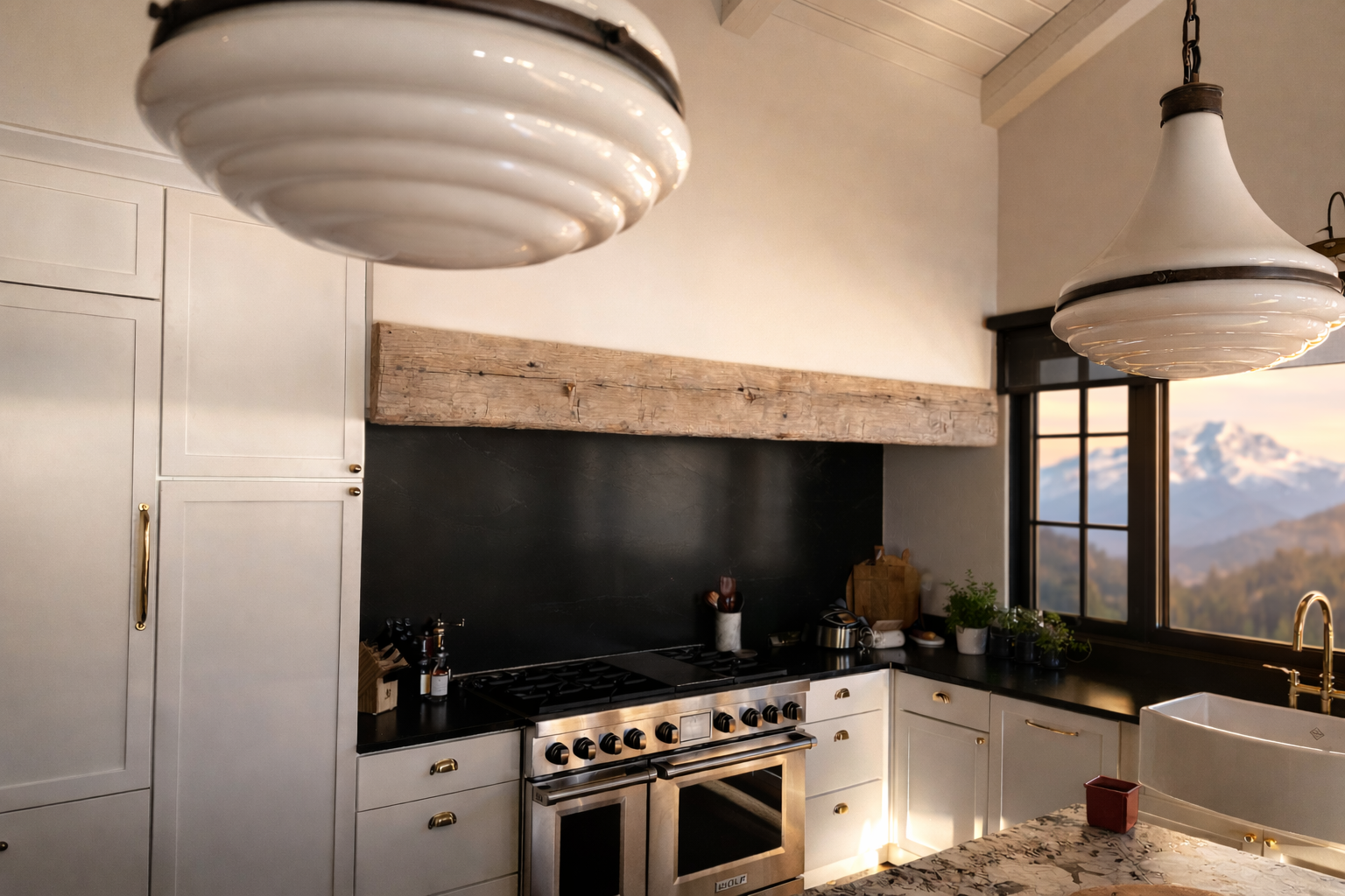 A modern kitchen with white cabinets, black countertops, a stainless steel stove, and large hanging pendant lights. There's a wooden beam above the stove and a window showing mountains outside.