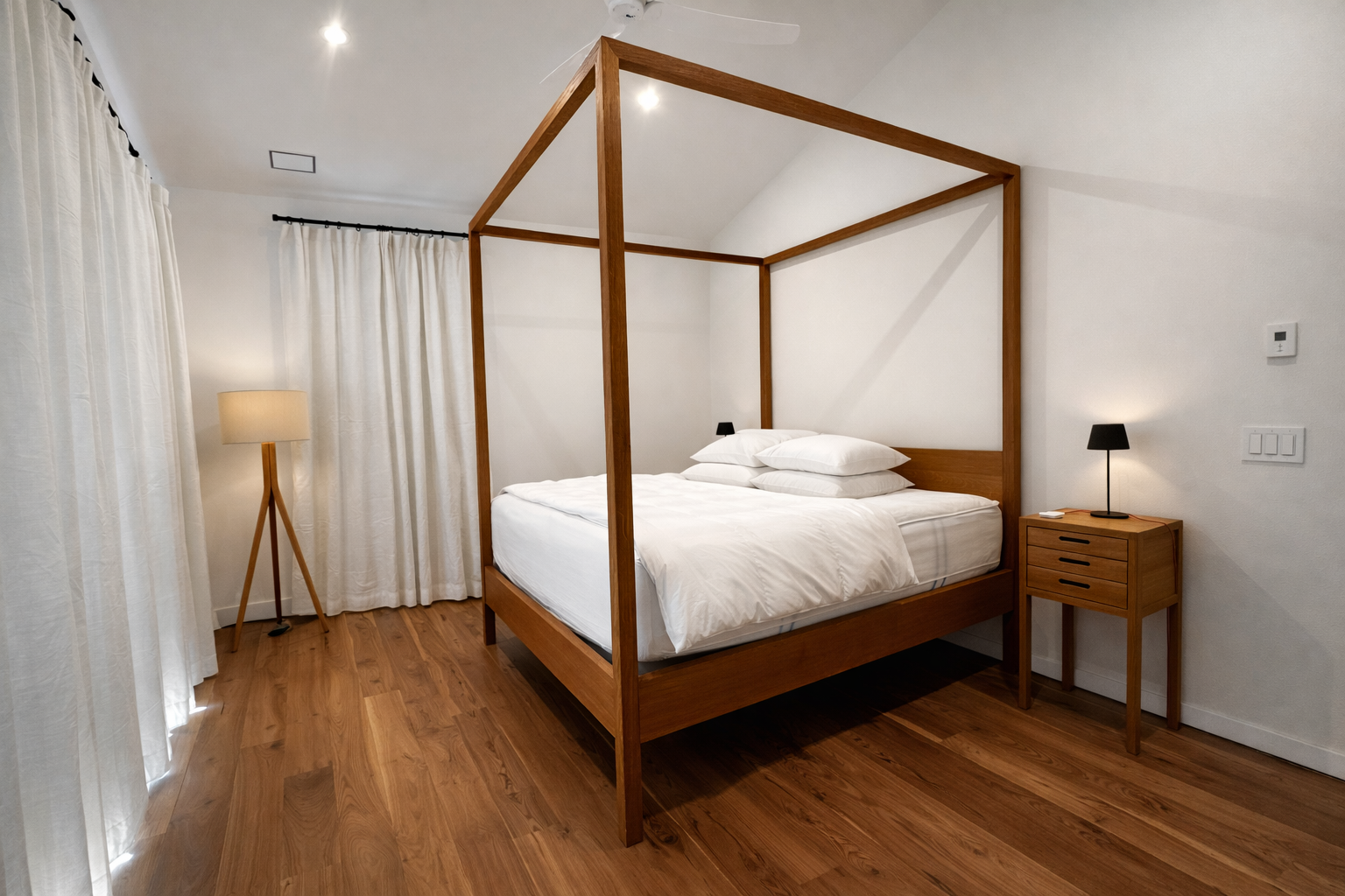 A bedroom with a four-poster wooden bed, white bedding, and four pillows. There is a wooden nightstand with a small black lamp on it, and a tall floor lamp with a tripod base. White curtains cover the window, and the floor is made of wood.