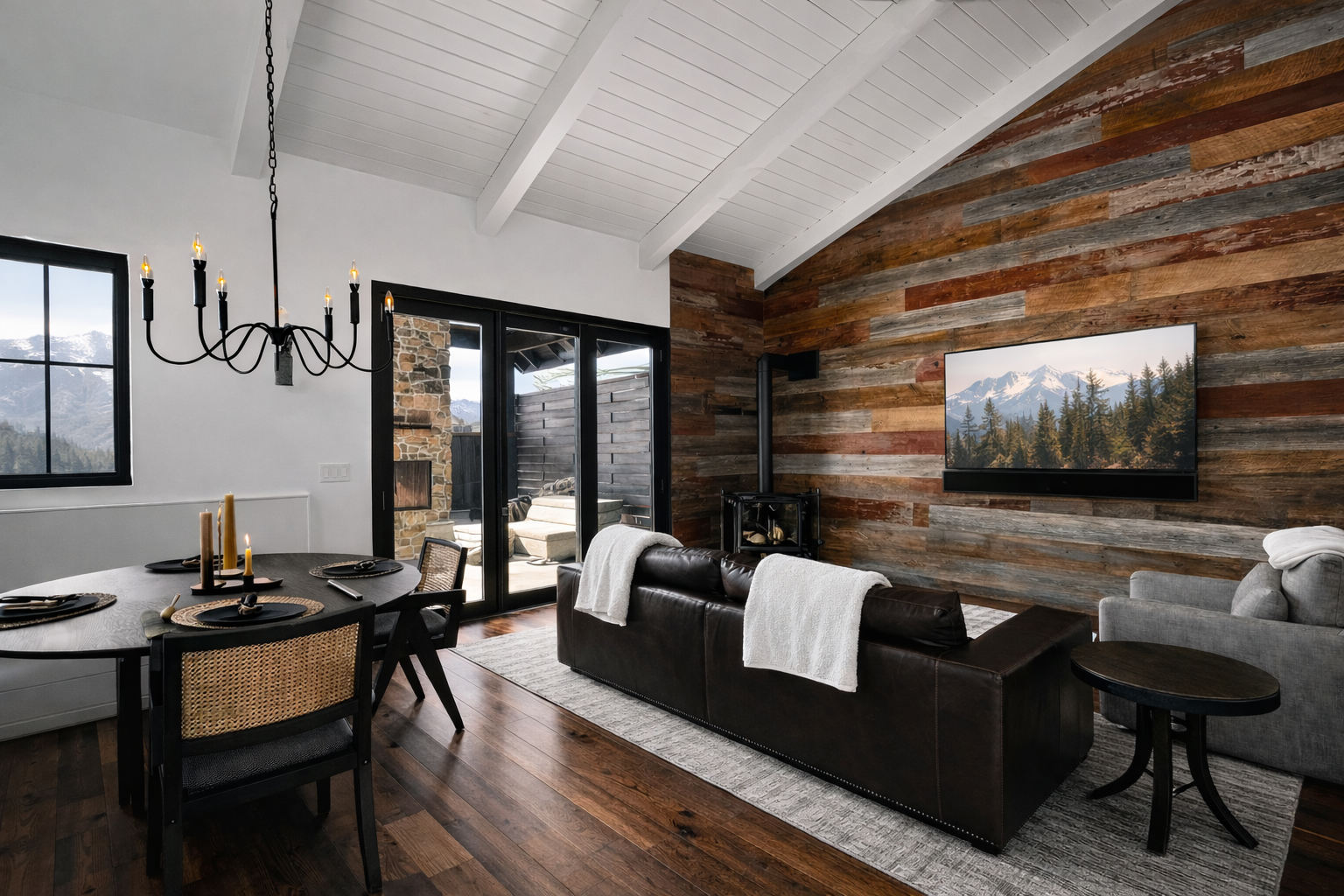 Living room with a wooden accent wall, a mounted flat-screen TV displaying landscape, a wood stove, dark leather sofa with white throws, a round dining table with black chairs, a chandelier, and large sliding glass doors leading outside to mountain v