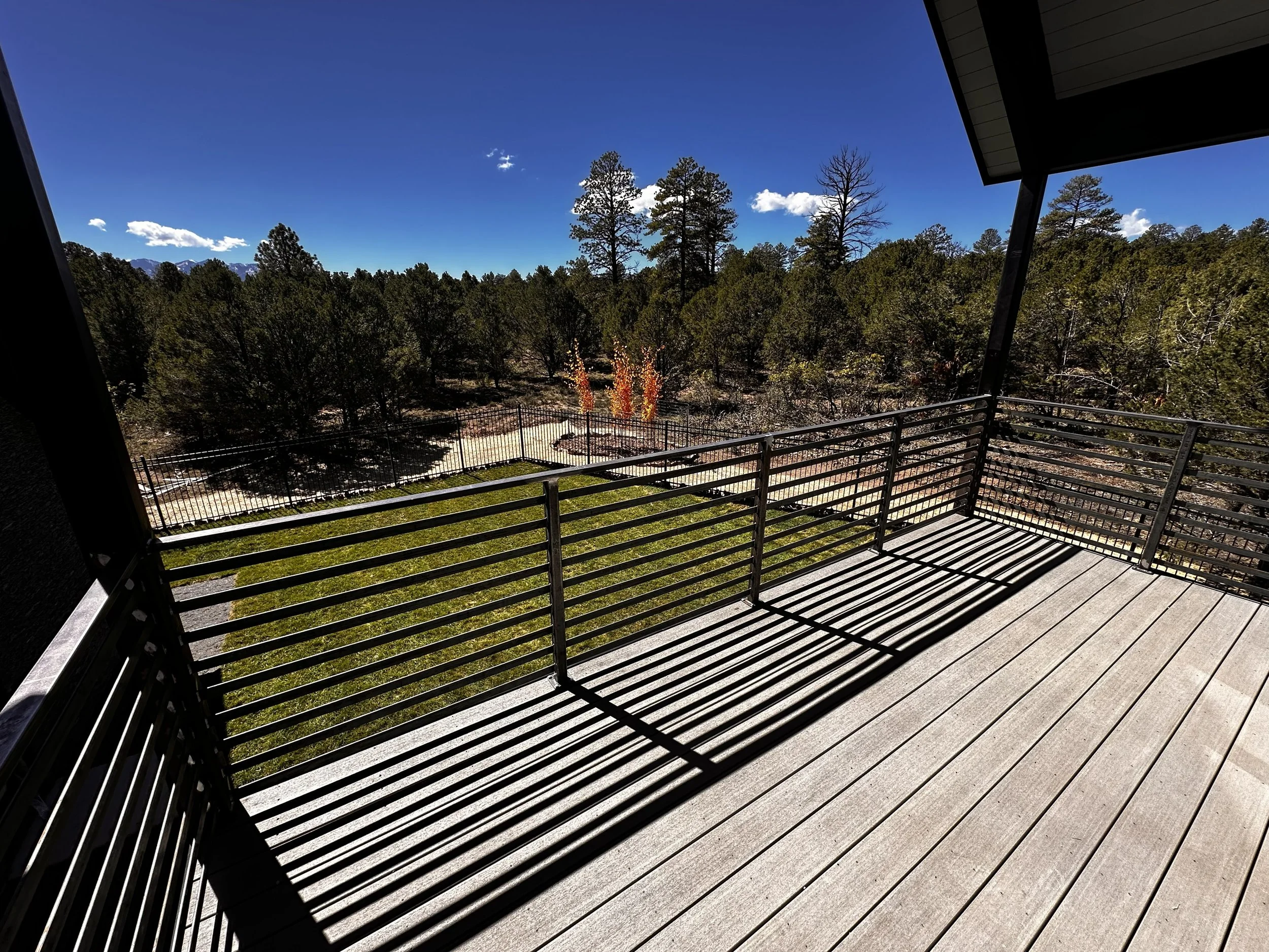 View from a wooden balcony overlooking a landscape with trees and a clear blue sky, with shadows cast by the railing.