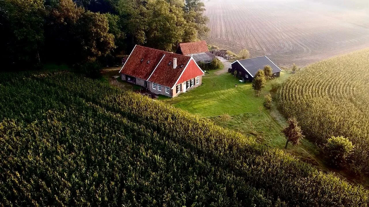 Een panoramisch uitzicht op een boerderij met meerdere gebouwen, omgeven door akkers en bossen, tijdens zonsondergang.