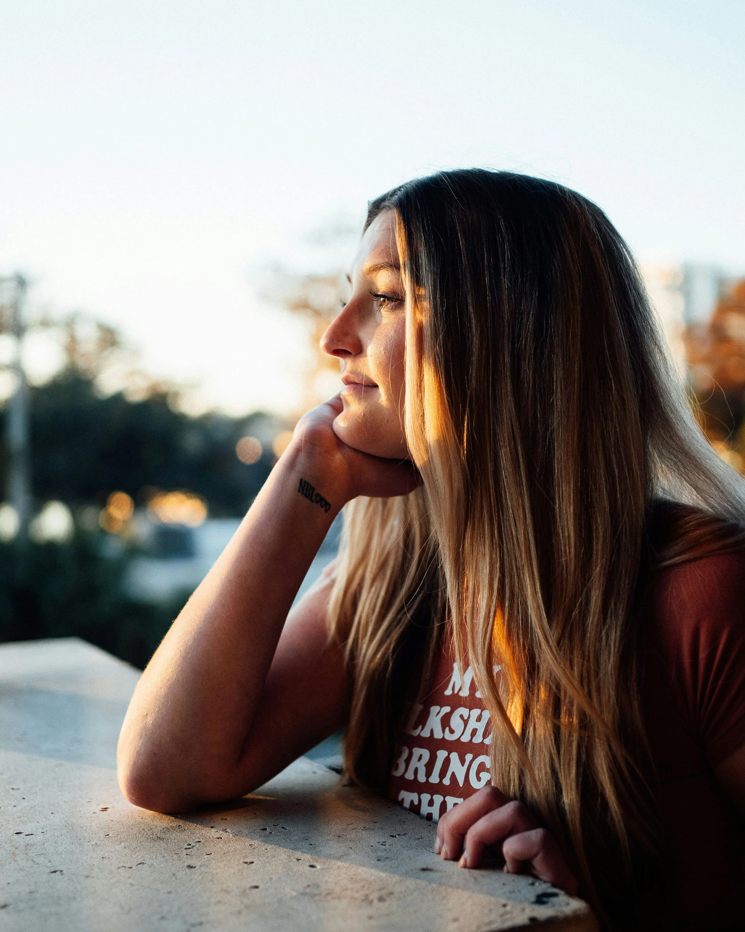 A woman with long hair sitting at a table, looking pensively towards a sunset.