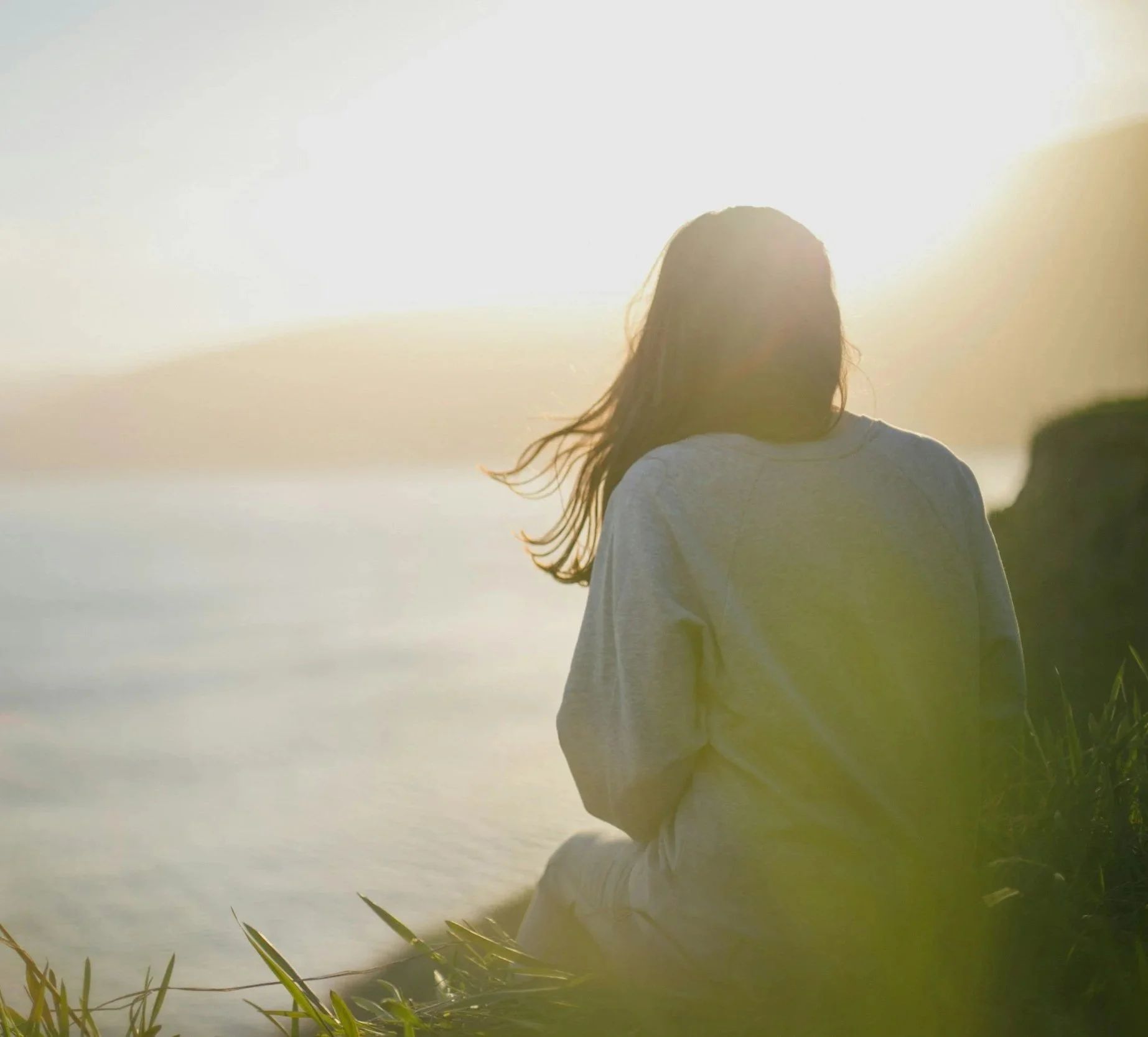 A person with long hair wearing a gray hoodie sitting on the grass near a body of water with the sun shining brightly in the sky.