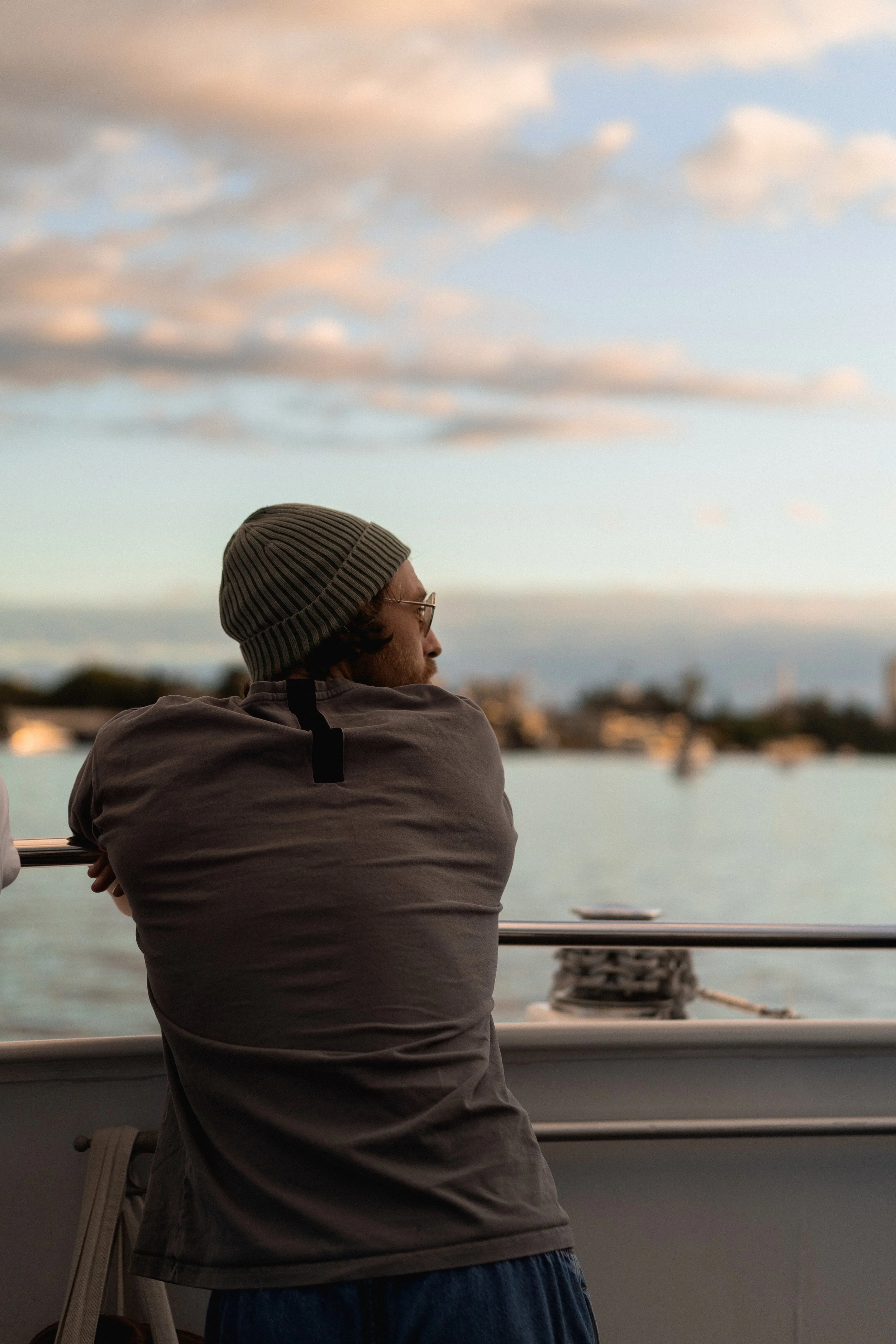 A man in a beanie and gray shirt looking out over a waterfront at sunset