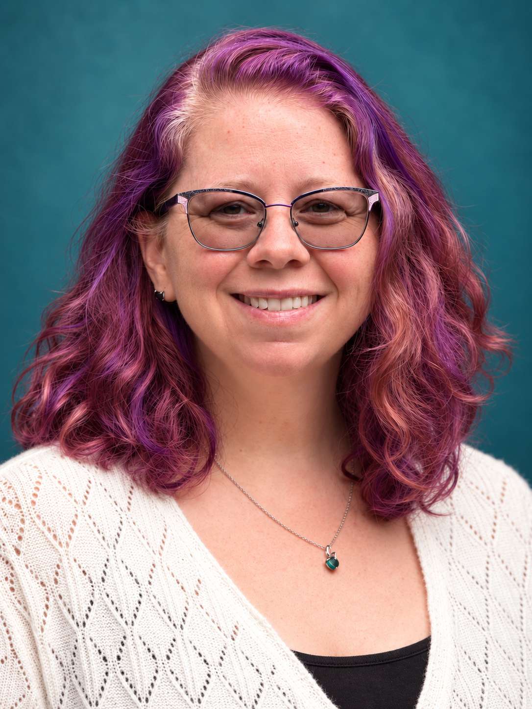 Close-up portrait of a woman with curly purple and pink hair, wearing glasses and a white knitted sweater, smiling against a teal background.