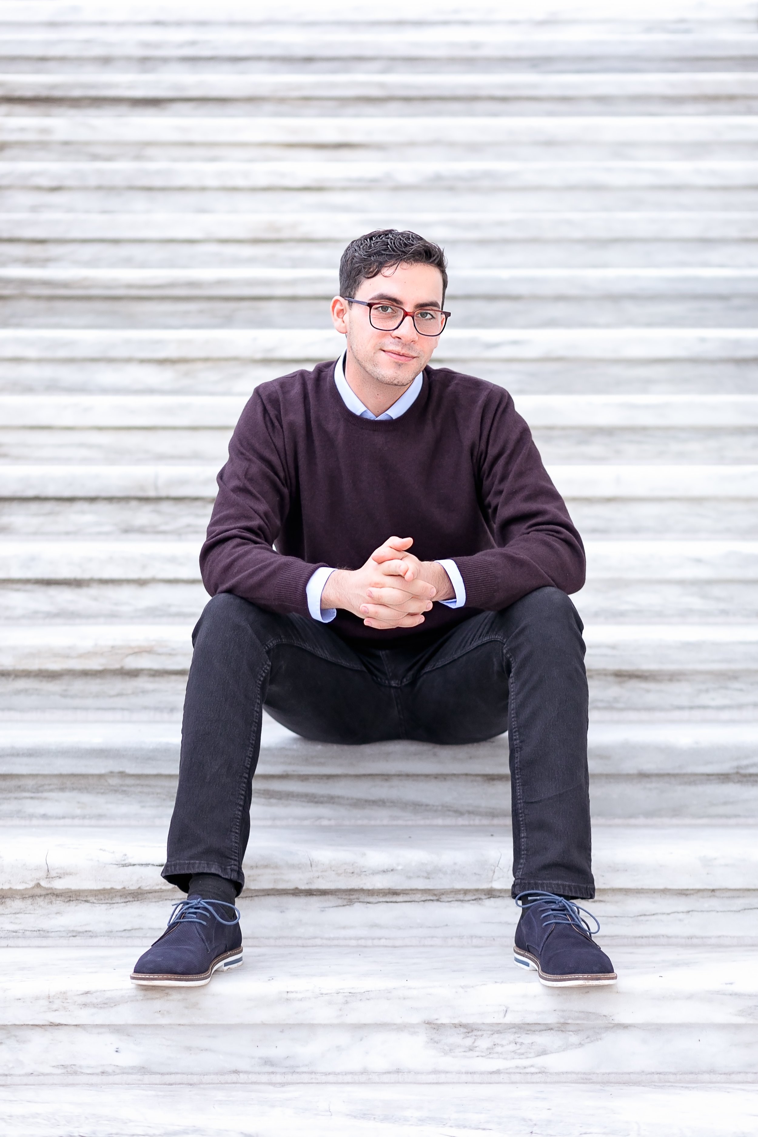 Man with glasses, dark sweater, and dark pants sitting on marble stairs.