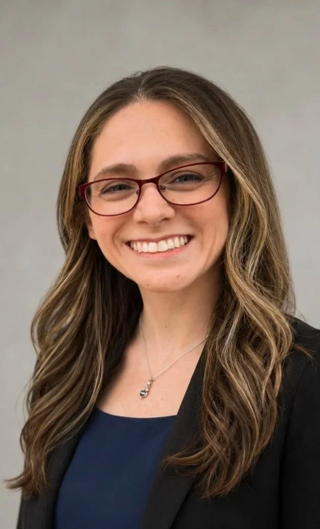 A smiling woman with long wavy brown hair, wearing red glasses, a dark blazer, a blue top, and a necklace, standing against a plain gray background.