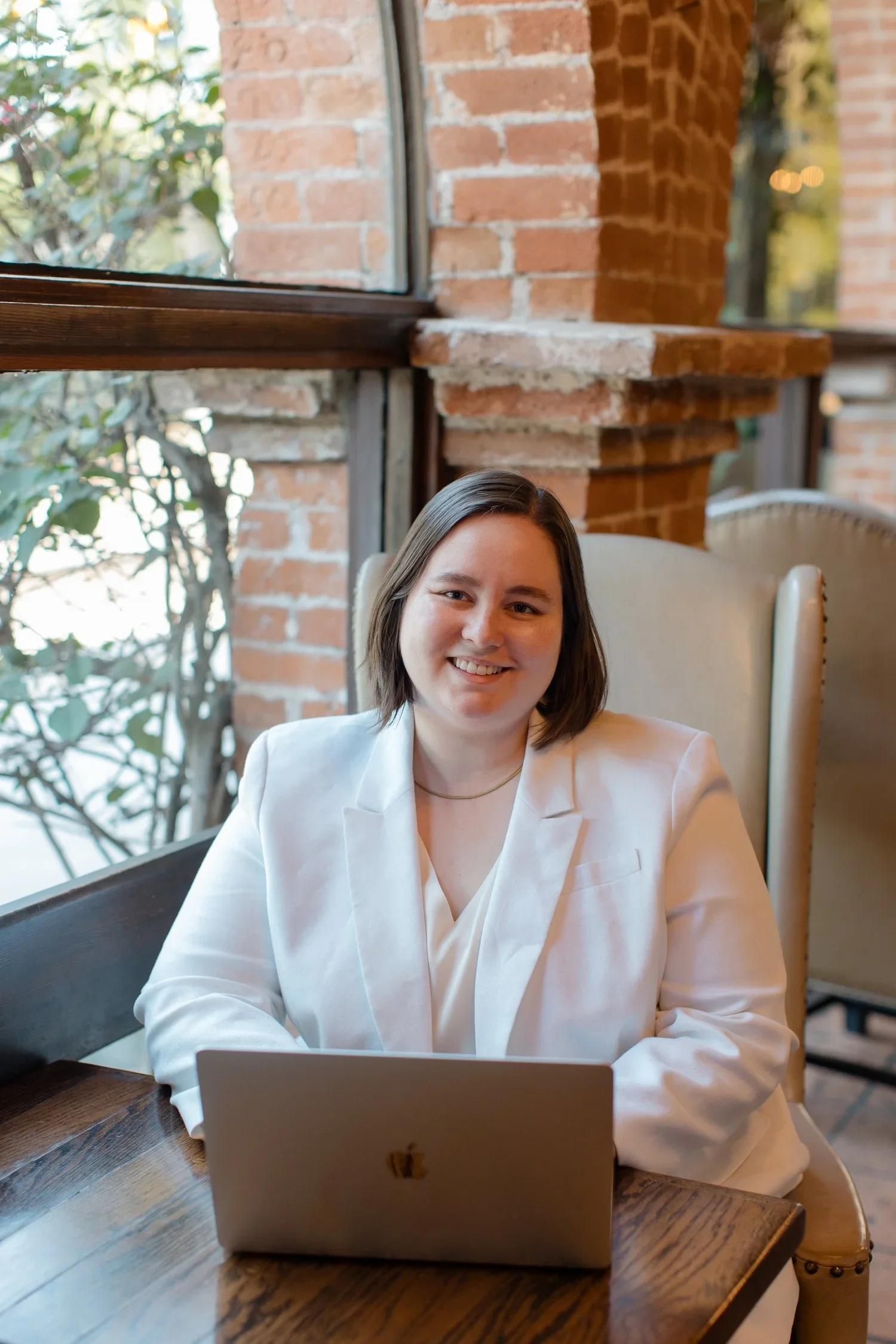 A woman with shoulder-length brown hair smiling, sitting at a wooden table with a silver laptop, in a cafe with brick walls and large windows.