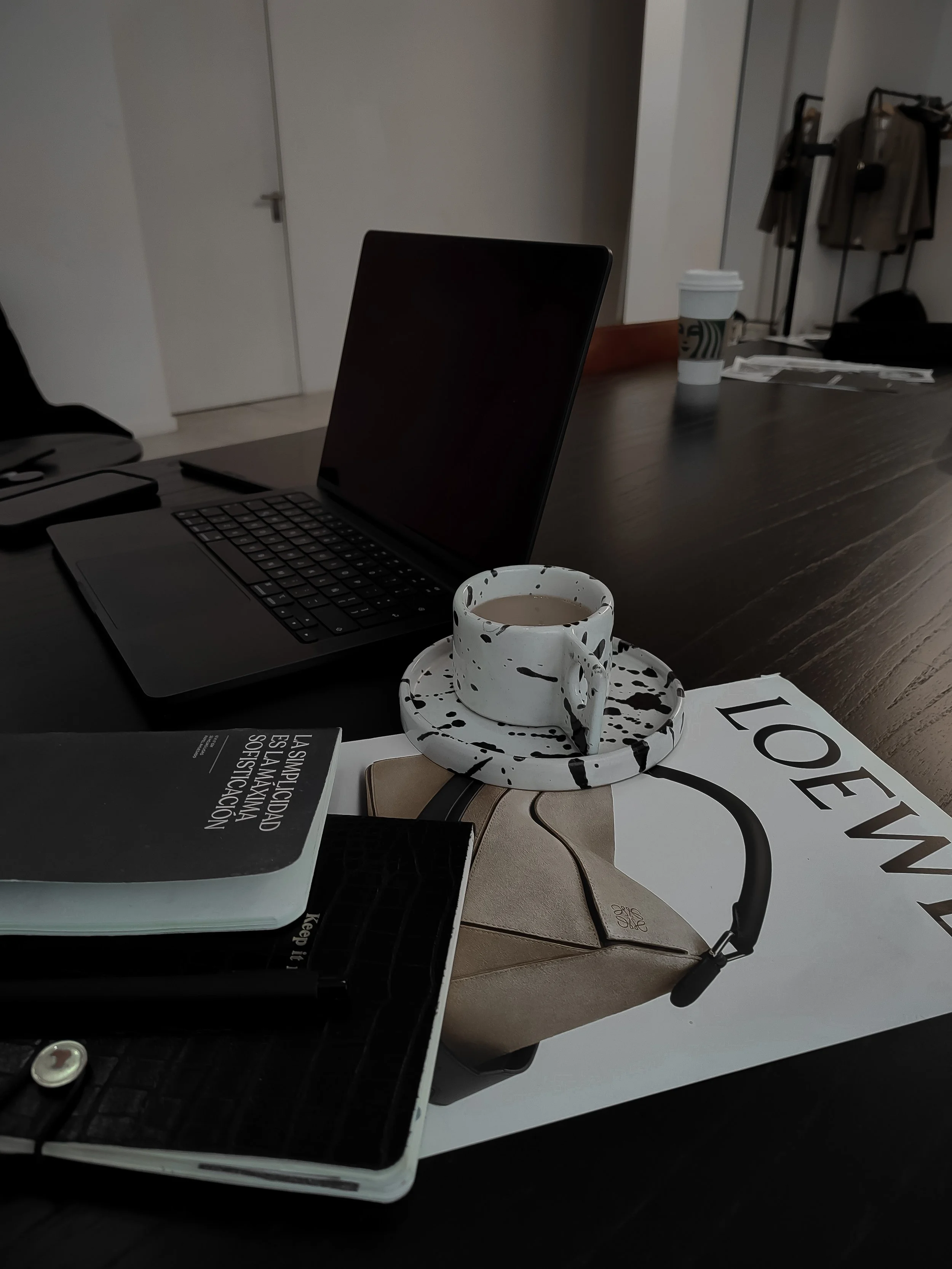 Black laptop, white speckled cup with coffee on a matching saucer, a beige handbag, magazines, and a Starbucks cup on a wooden desk, with a doorway and clothes in the background.