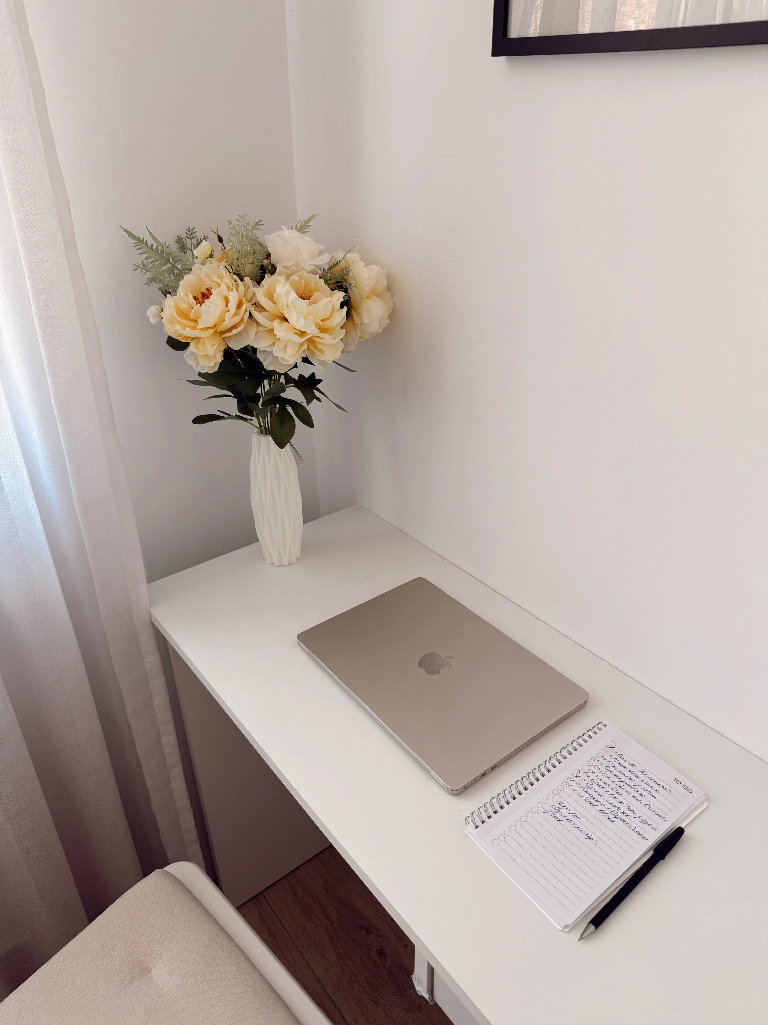 A white desk with a closed silver Apple MacBook, a notebook with handwritten notes, a black pen, and a tall white vase with cream-colored flowers and green foliage inside.