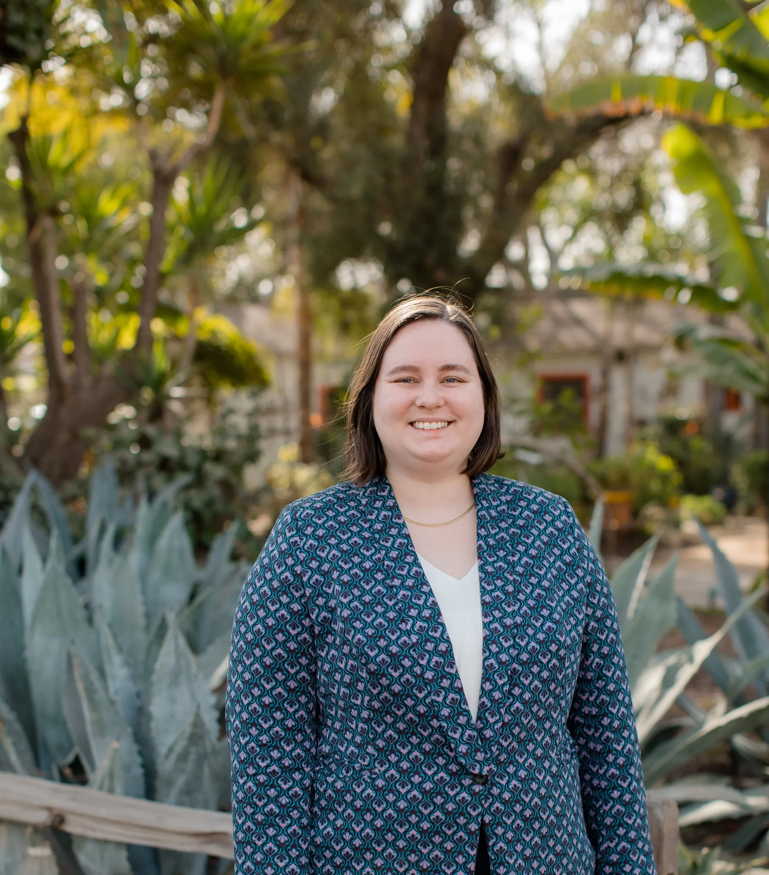 A woman smiling outdoors in front of trees and plants during the daytime.