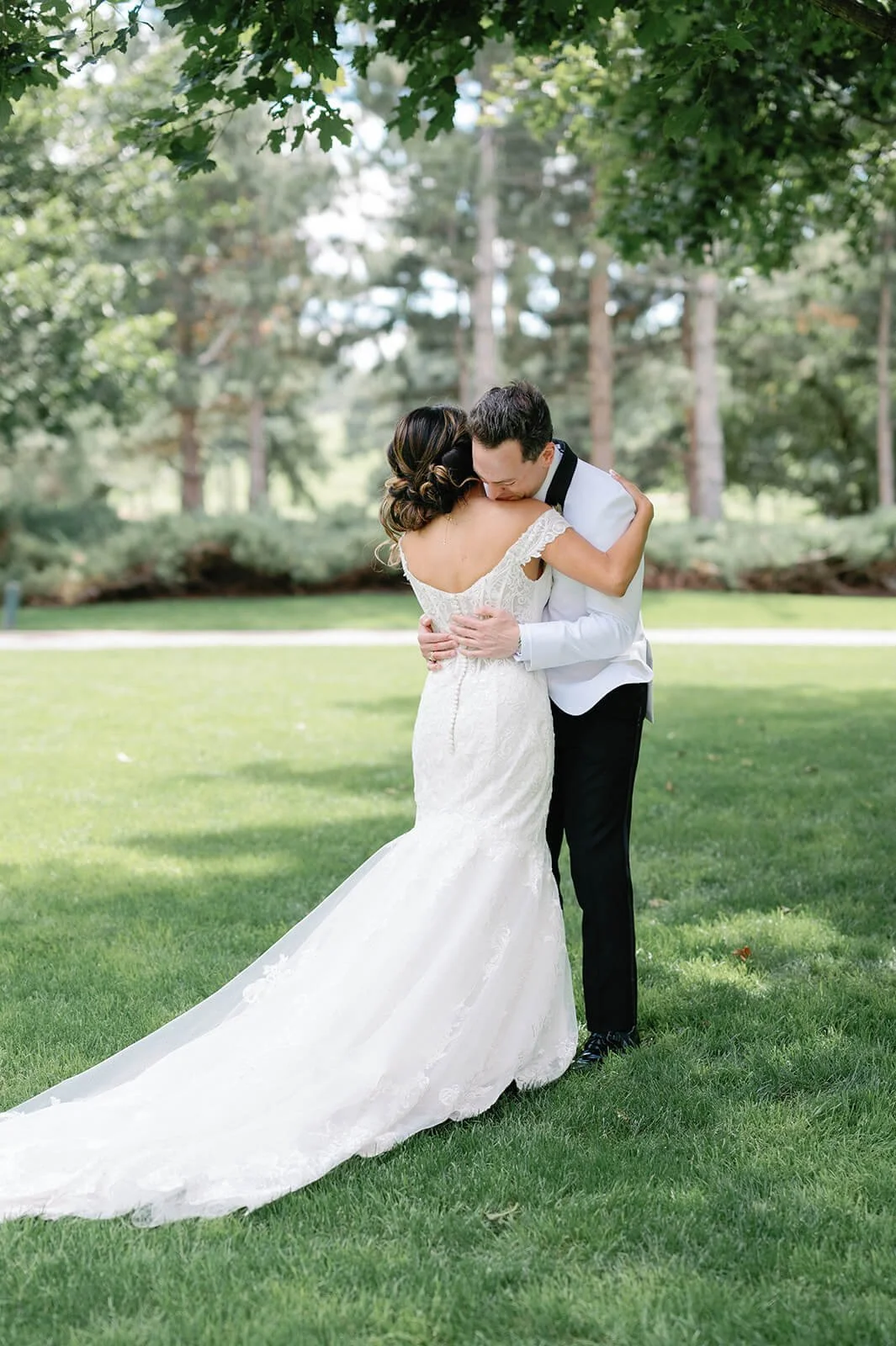 A bride and groom embracing each other outdoors on a grassy area in a park with trees in the background, during a wedding celebration.