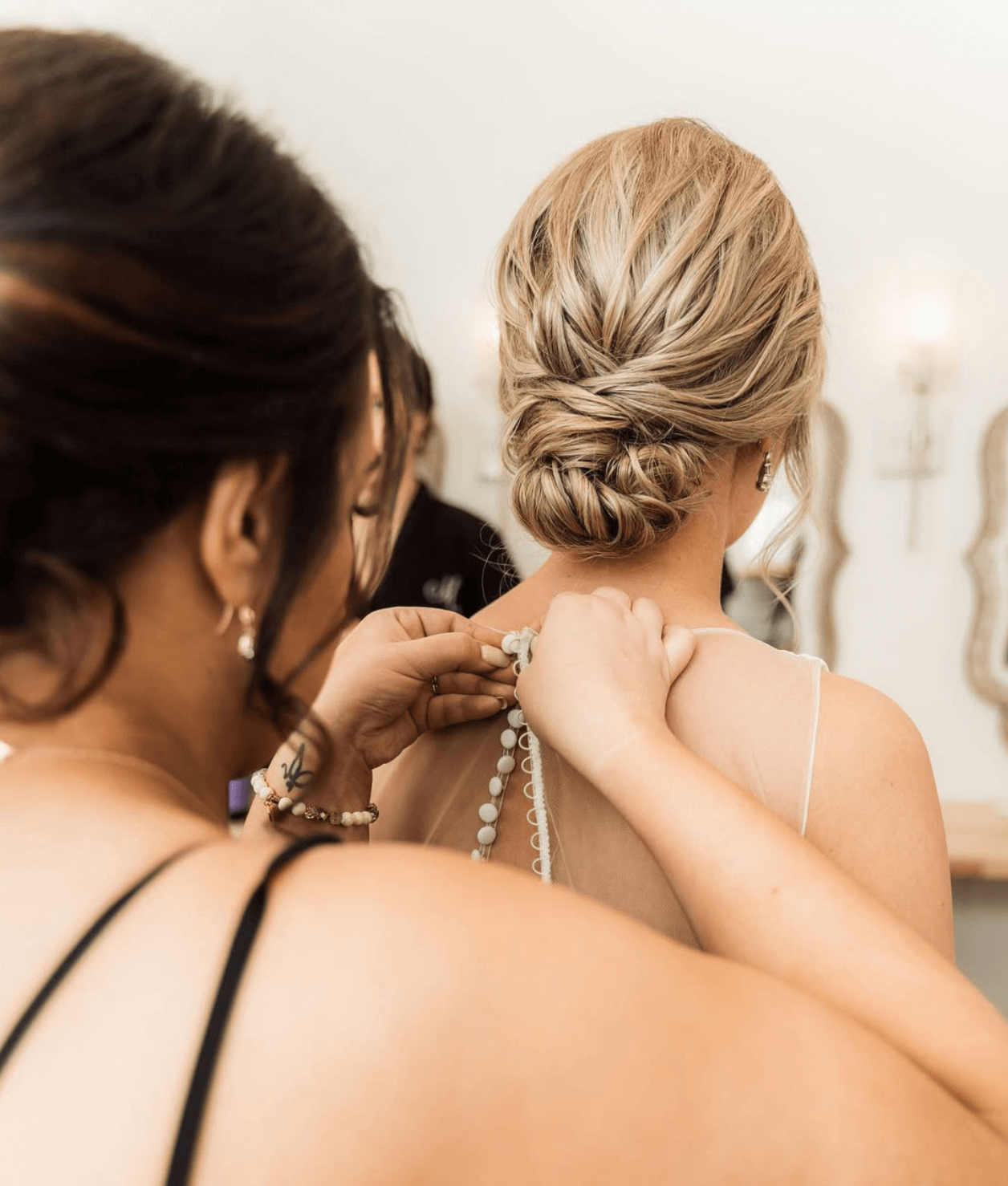 A woman wearing a wedding dress is being assisted with her necklace by another woman with dark hair, in a bridal preparation setting.