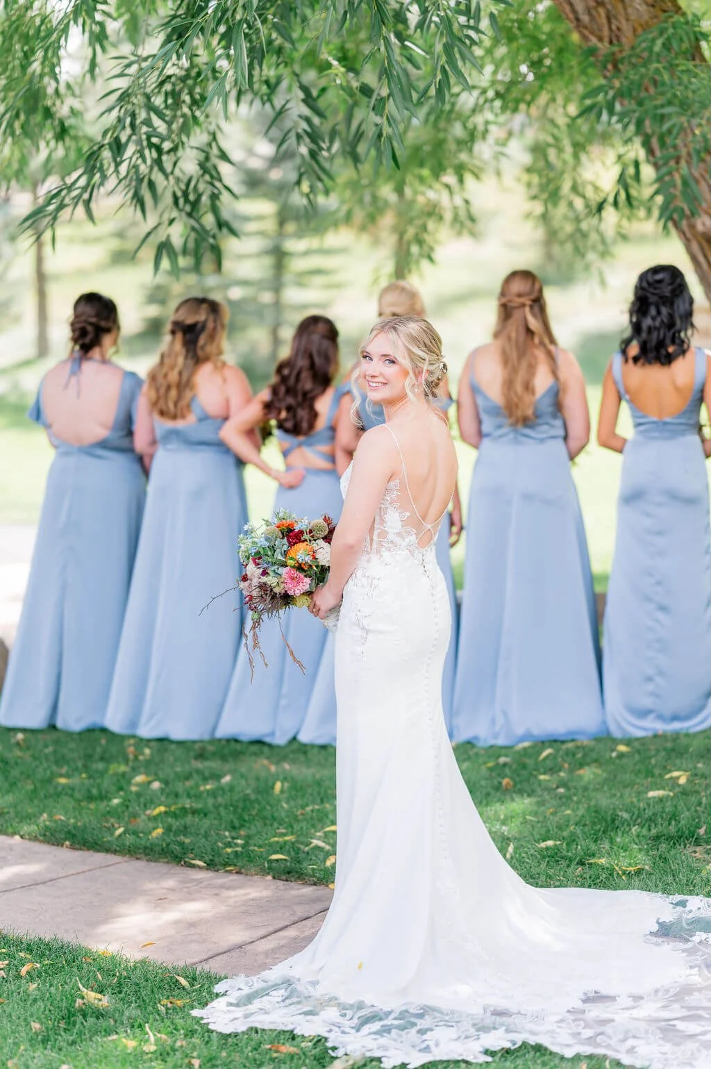 A bride holding a bouquet of flowers in front of her, with six bridesmaids standing behind her under a large tree in an outdoor setting during daytime.
