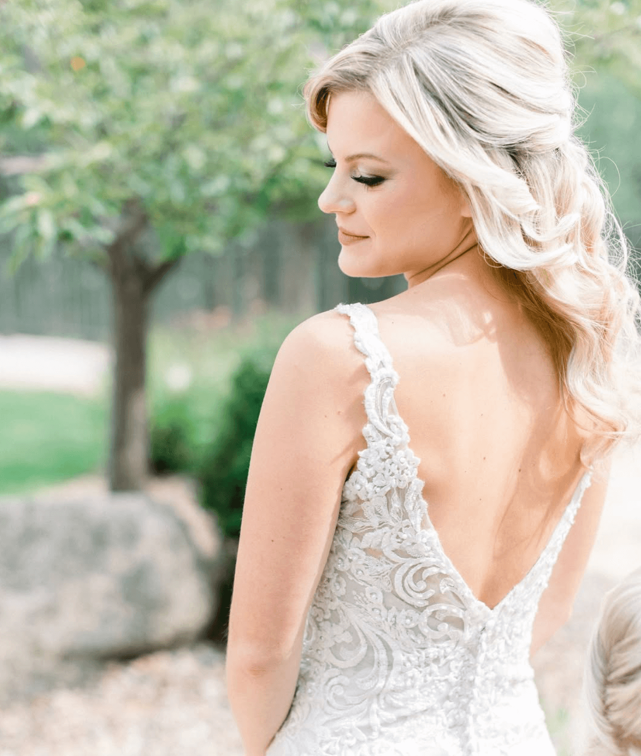 A bride with blonde hair styled in loose waves, wearing a white lace wedding dress with exposed shoulders, standing outdoors with a blurred background of trees and greenery.