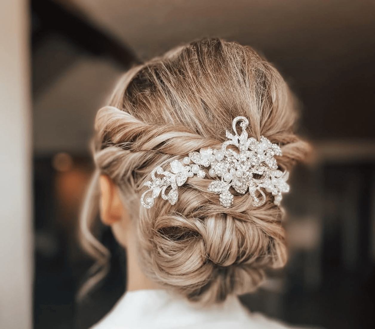 Back view of a woman with an elegant braided updo hairstyle, adorned with a large, ornate silver hairpiece decorated with pearls and crystals.