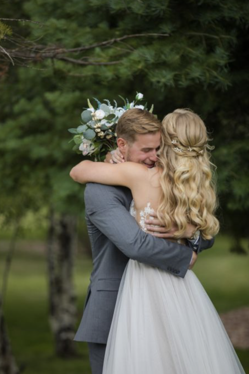 A bride and groom embrace outdoors in a romantic wedding scene, with the bride holding a bouquet of white and green flowers. The groom is wearing a gray suit, and the bride has long, wavy blonde hair adorned with a hairpiece.