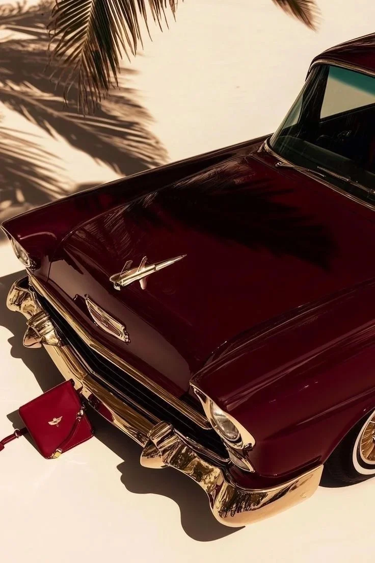 A vintage maroon car with chrome accents parked on a white surface, with a red handbag placed in front of it. A palm tree's shadow is cast on the ground nearby.
