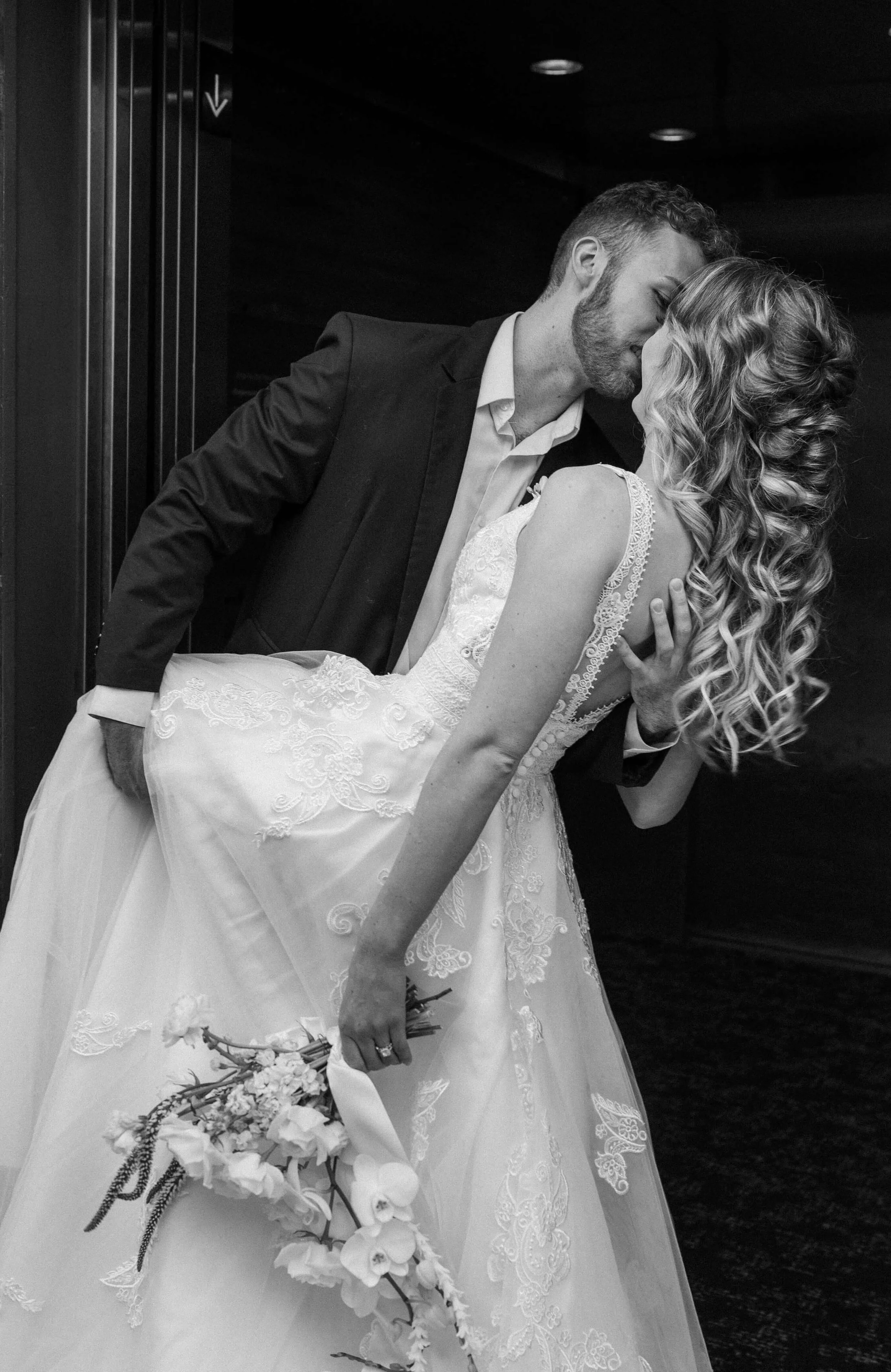 A black and white photo of a newlywed couple sharing a kiss in an elevator. The groom is dressed in a suit, and the bride is in a lace wedding gown, holding a bouquet of flowers.