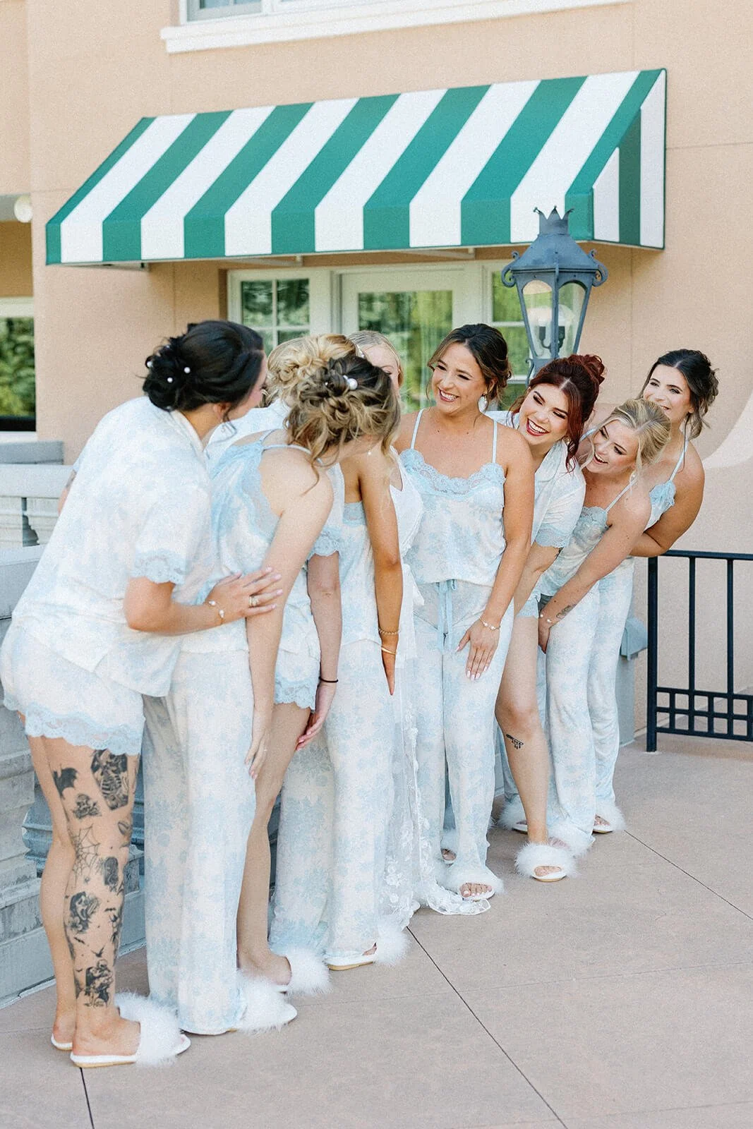 Group of women wearing pajamas, standing outside a building with a green and white striped awning, laughing and chatting.