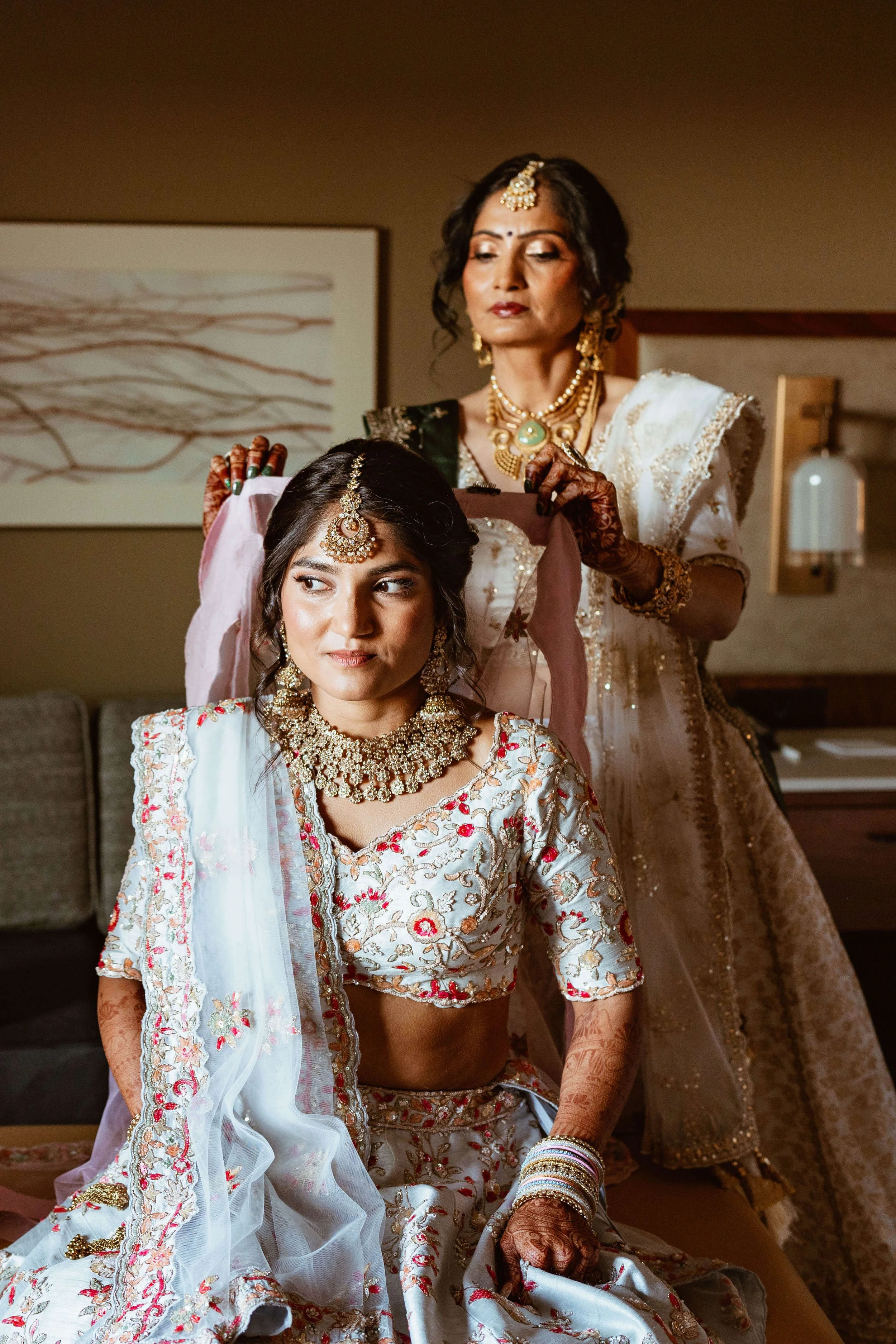 Two women dressed in traditional Indian bridal attire, one sitting and the other standing behind her, adjusting her jewelry in a room with modern decor.