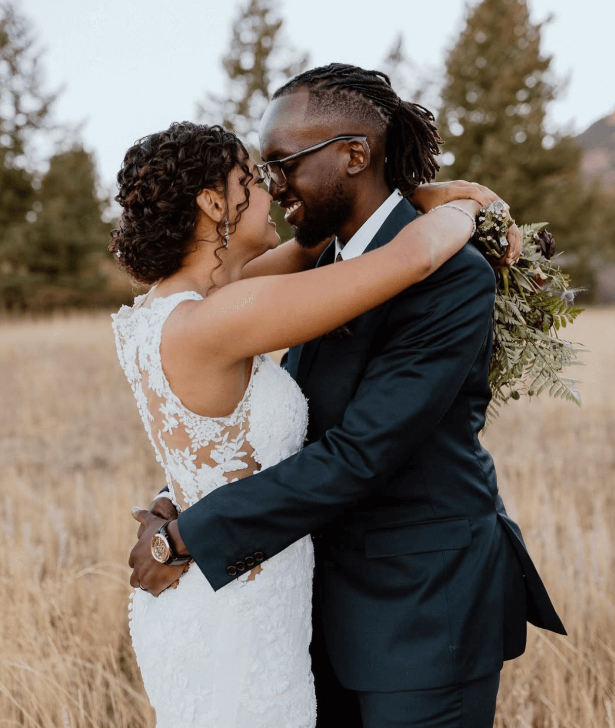 A newlywed couple embracing outdoors in a field with trees in the background.