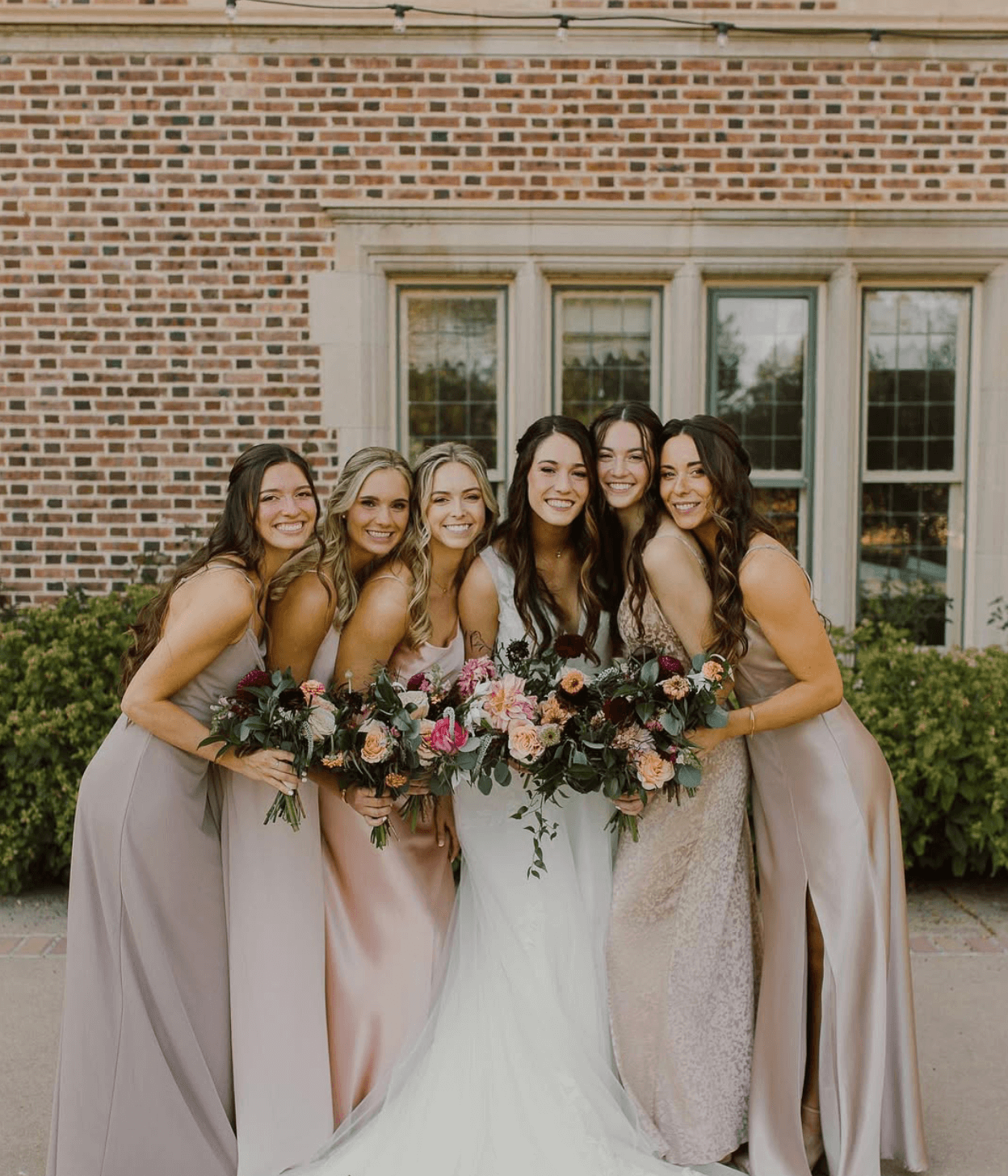 Group of six women in dresses standing outdoors in front of a brick building holding bouquets of flowers, smiling.