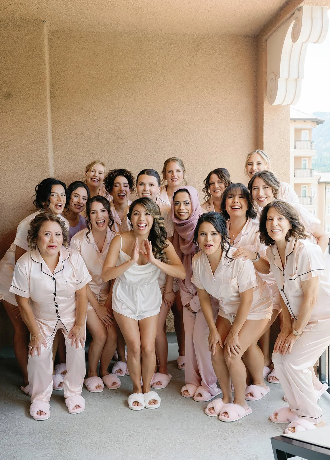 Group of women and a girl in pajamas celebrating on a balcony with beige walls, some wearing pink slippers, smiling and posing for the photo.