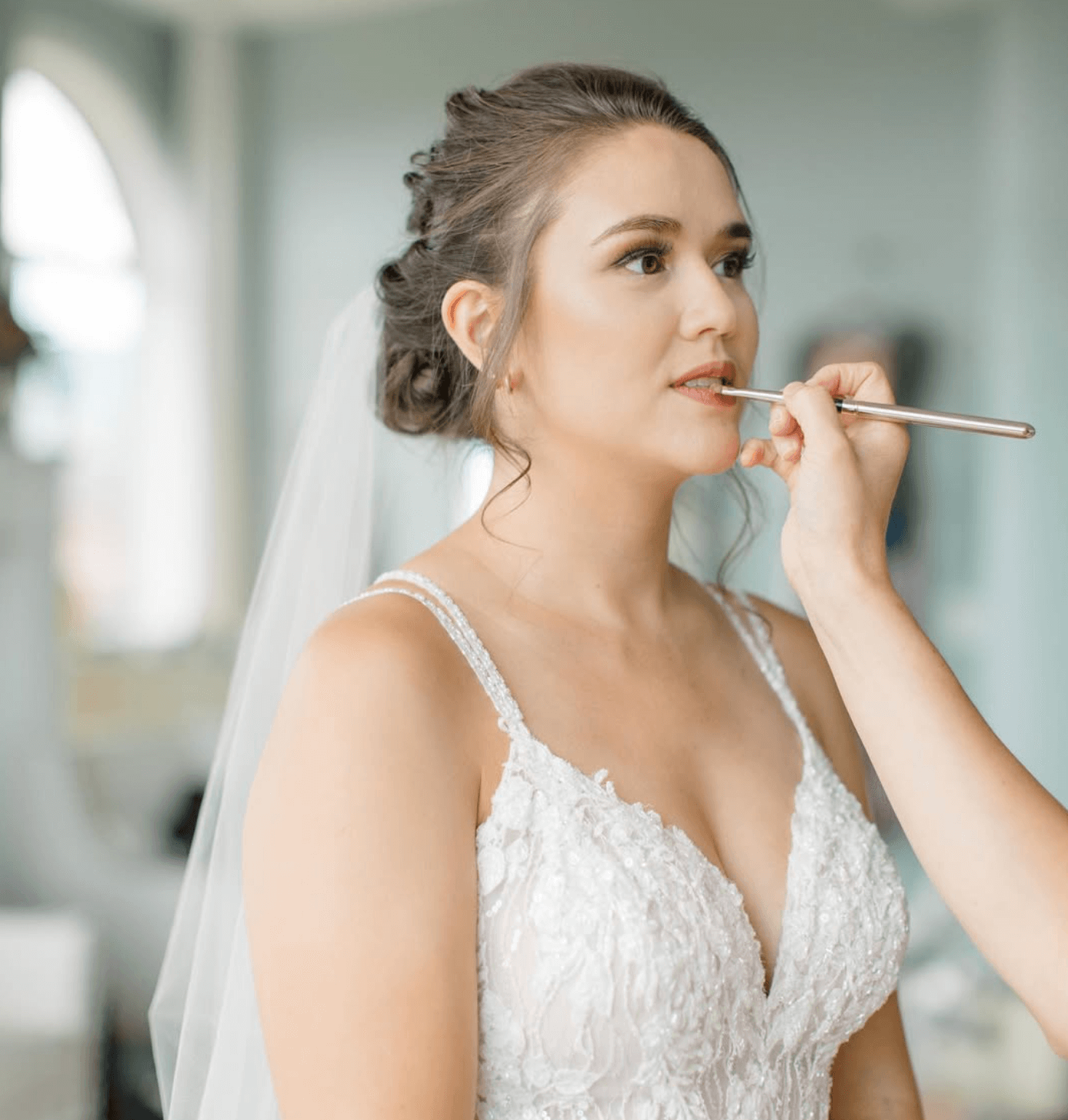 A bride getting her makeup done, wearing a white wedding dress with lace details and a veil, while a makeup artist applies lipstick with a brush.