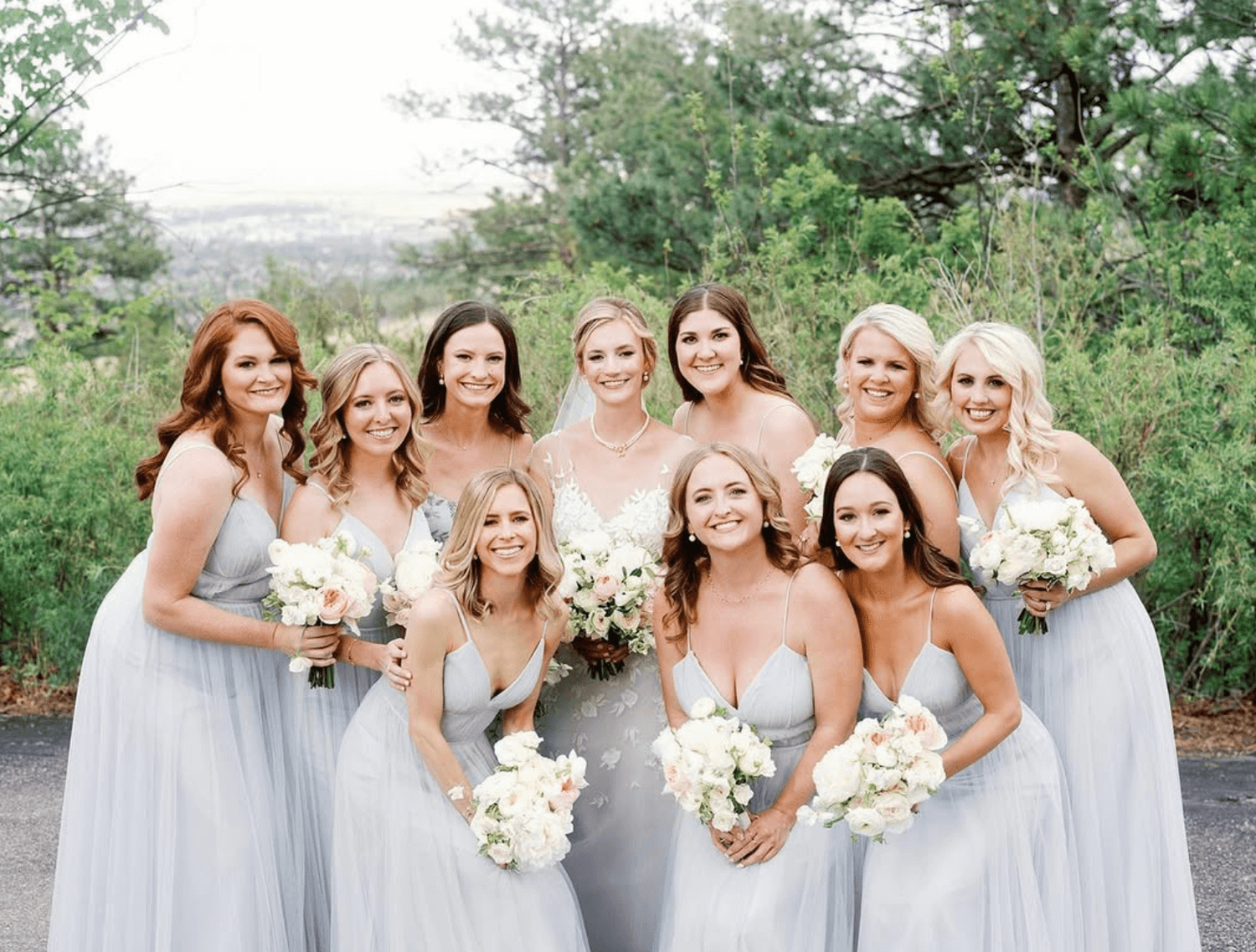 Group of ten women in light-colored dresses, posing outdoors with greenery in the background, smiling, holding bouquets of white and pink flowers.