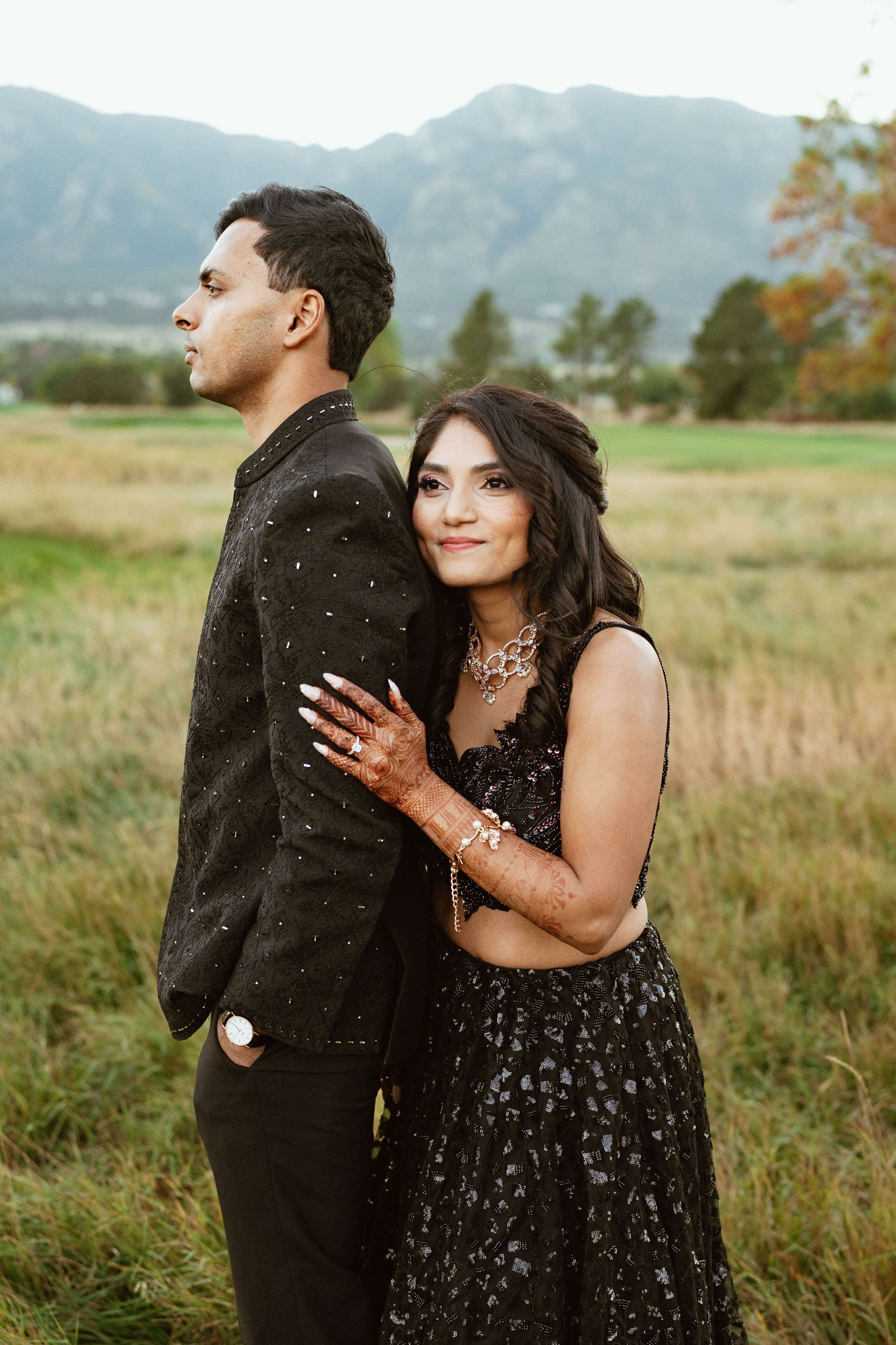A woman dressed in black with henna tattoos on her hands hugging a man in a black jacket in a grassy field with mountains in the background.