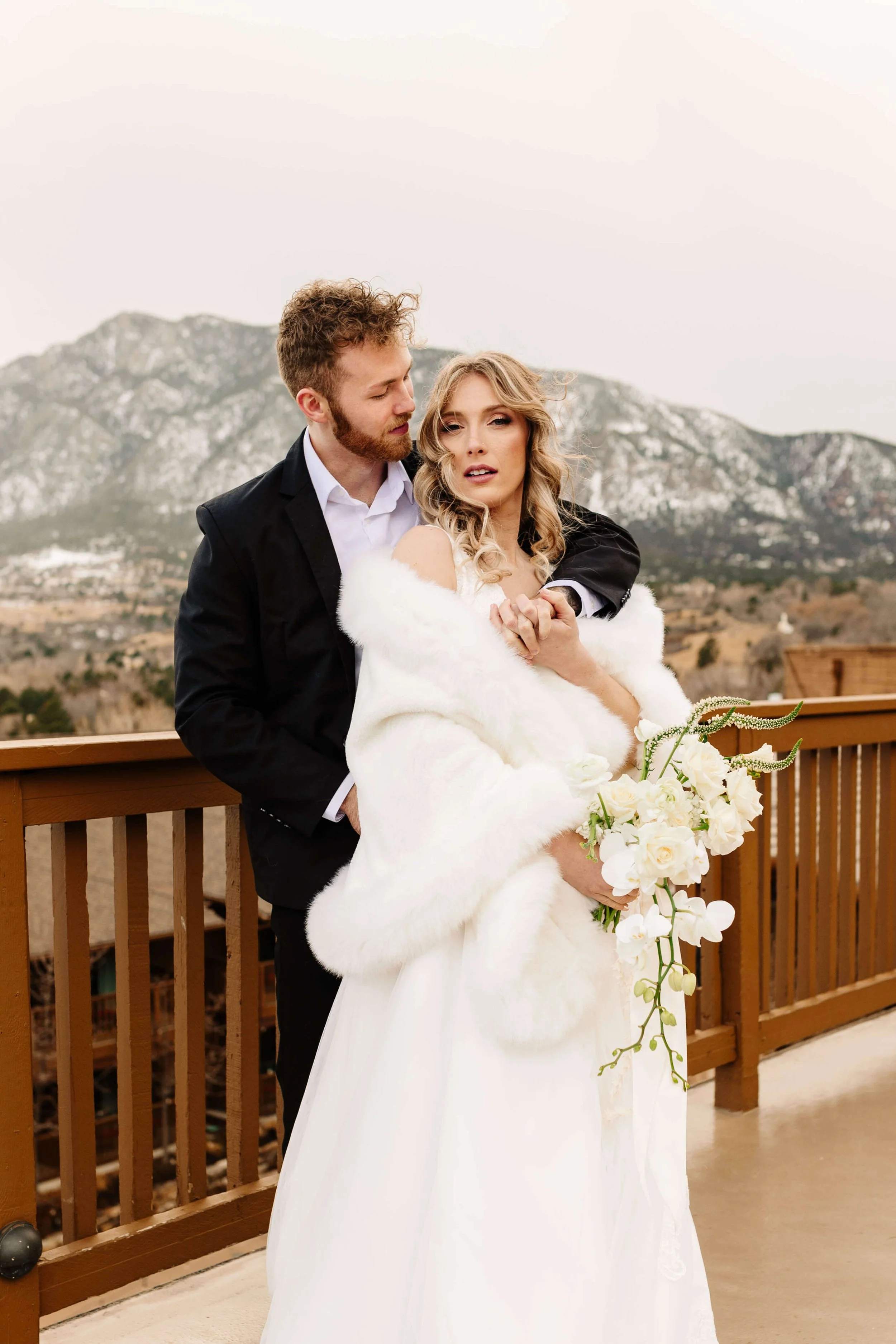 A bride and groom on a balcony with mountains in the background. The bride is holding a bouquet of white flowers and wearing a white dress with a faux fur stole. The groom is dressed in a black suit with a white shirt, embracing the bride.