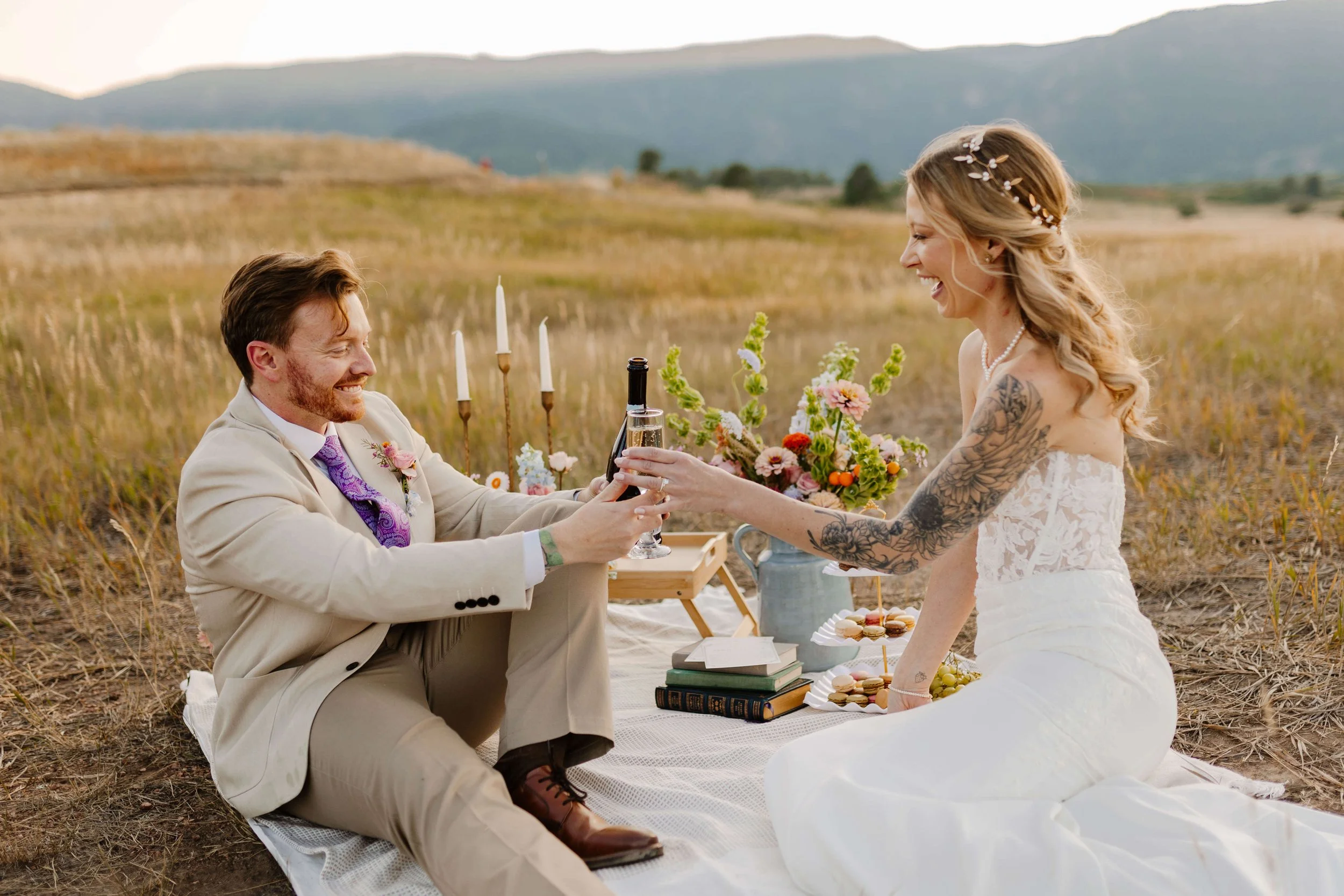 A bride and groom having a picnic-style wedding celebration in a field, sharing a toast with glasses of wine, surrounded by flowers and open landscape.