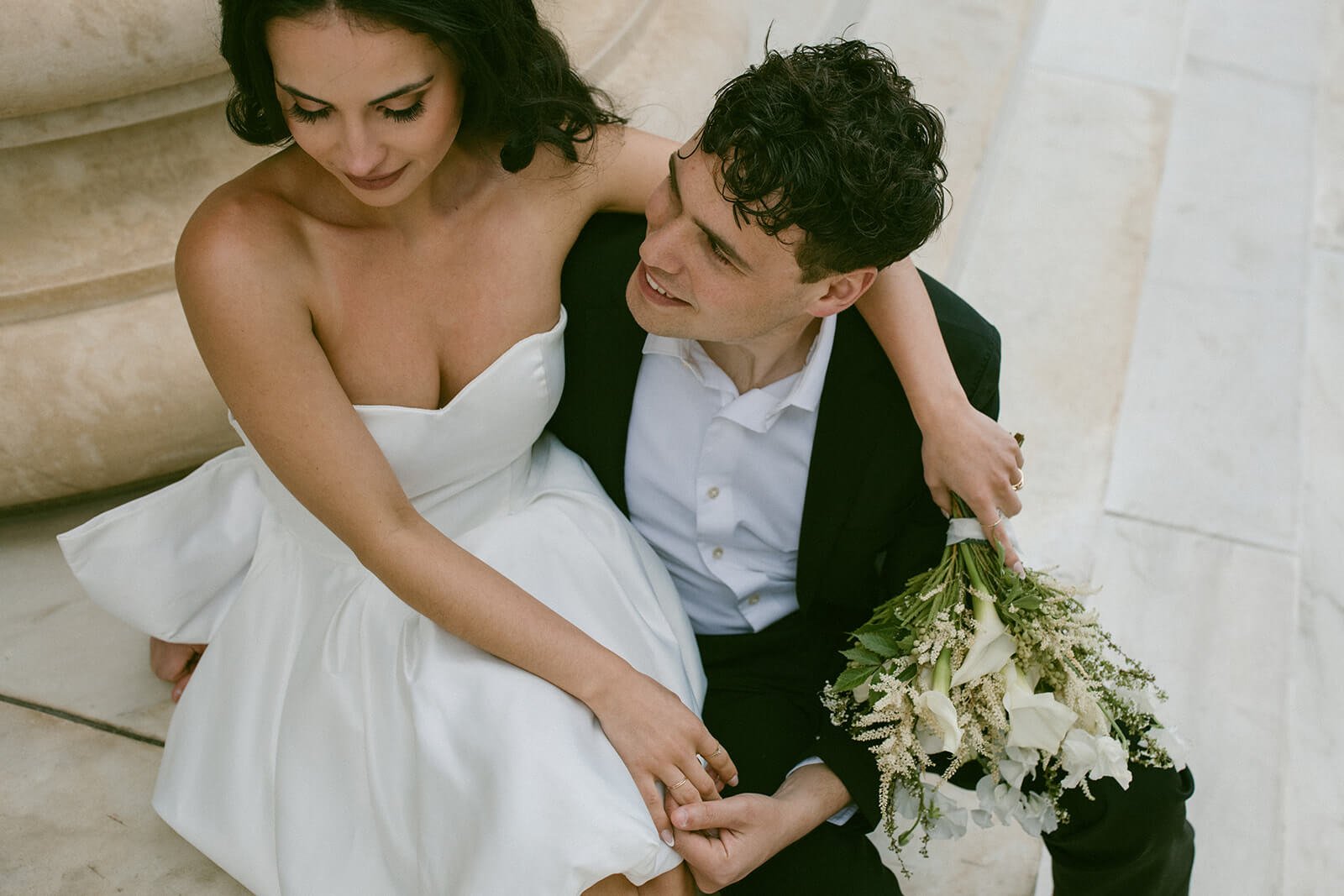 A woman in a white wedding dress sitting on a staircase, holding hands with a man in a suit who is sitting next to her, holding a bouquet of white flowers.