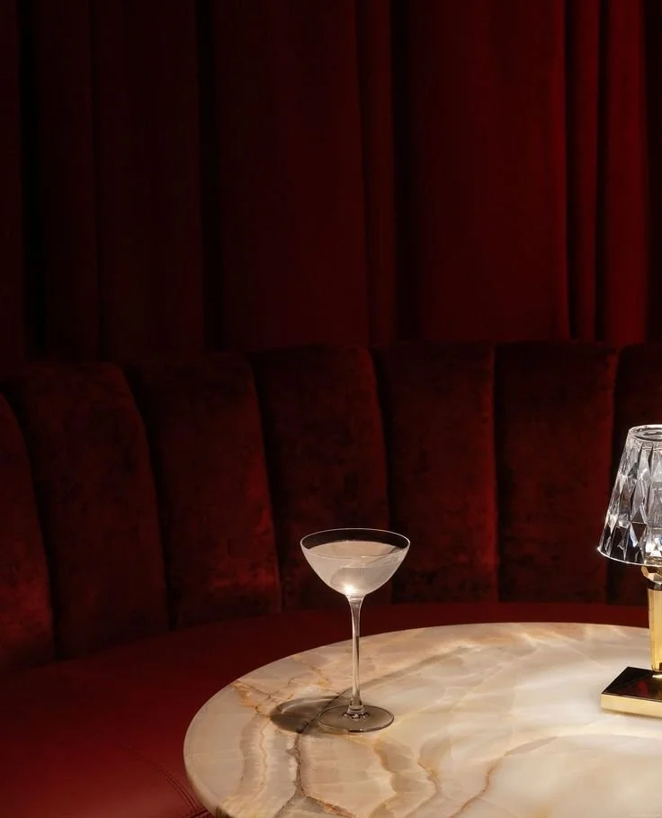 A cocktail glass filled with a clear drink on a round marble table, with a crystal lamp to the right.