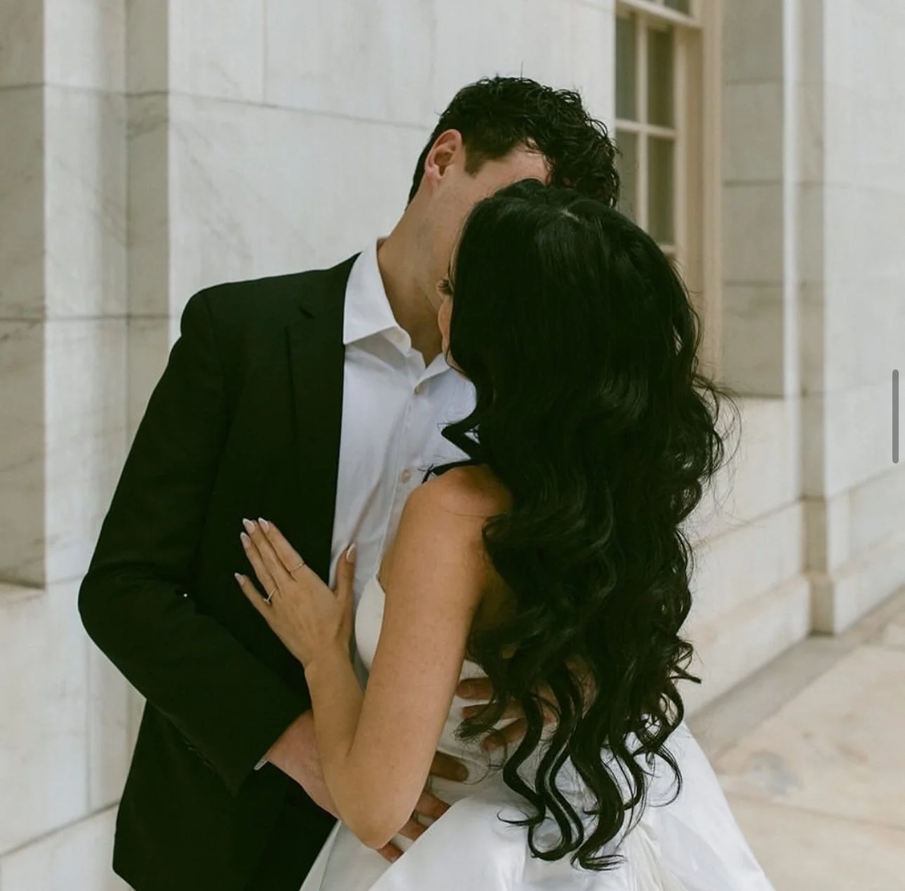 A bride and groom in formal attire sharing an intimate moment, with the man in a black suit and white shirt and the woman in a white dress with long black hair extensions, standing close to each other against a stone building background.