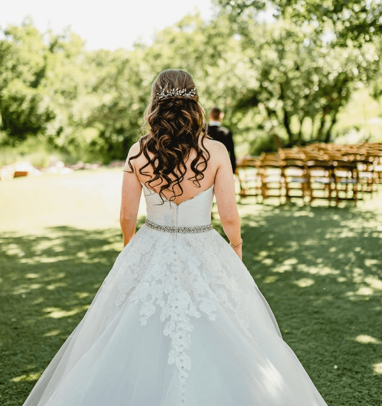 Bride in a strapless white wedding gown with lace details and a beaded belt, standing outdoors in a green garden with a man in the background.