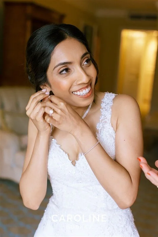 A woman in a white wedding dress putting on earrings, smiling happily.