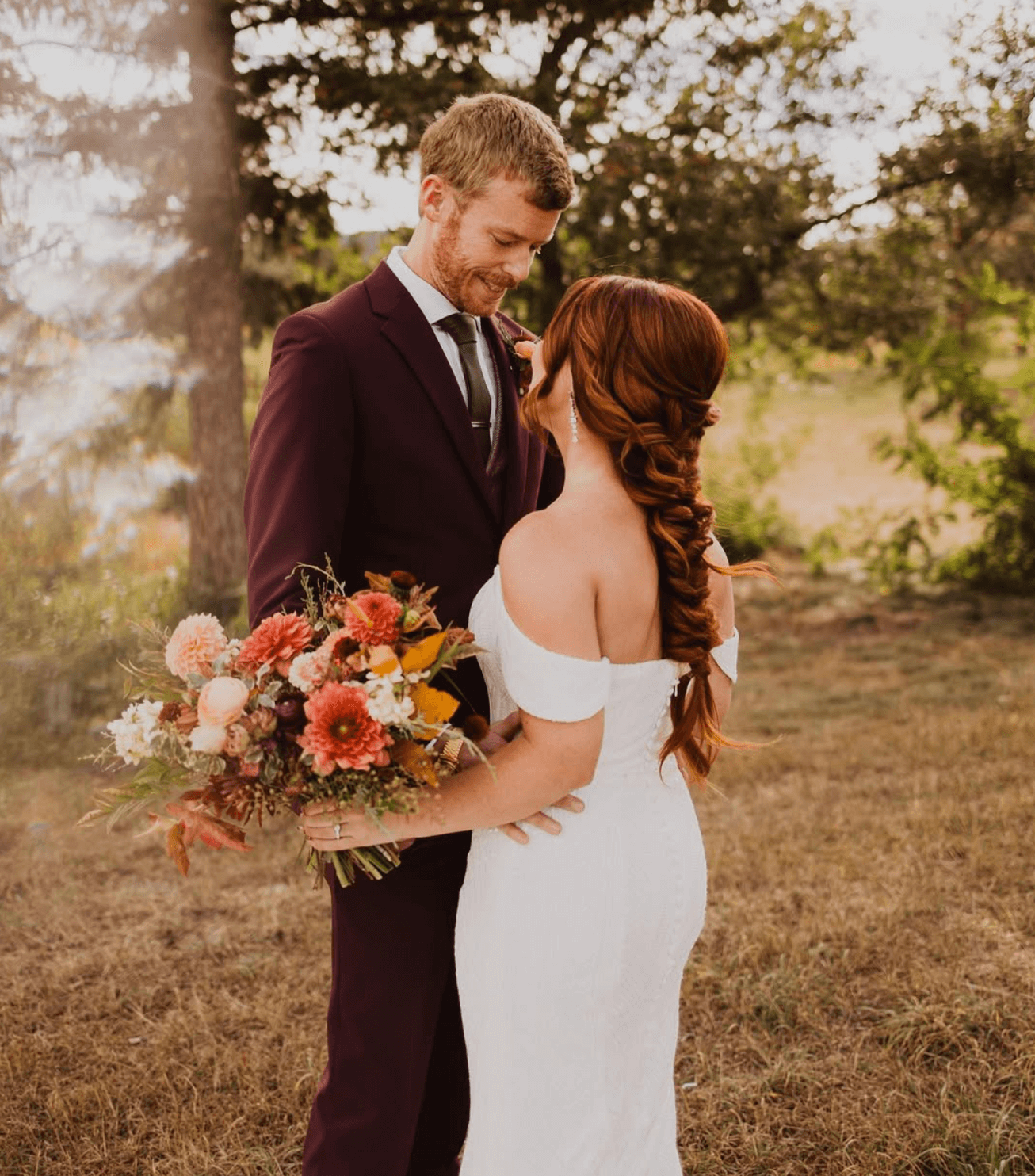 A man in a tuxedo and a woman in a white wedding dress stand close together outdoors, with the man holding a bouquet of pink, orange, and white flowers. They are in a natural setting with trees and grass.