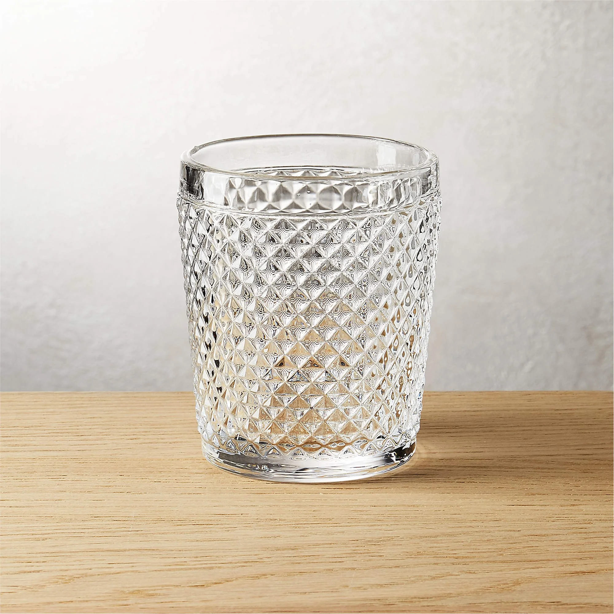 A clear, textured glass tumbler with a diamond pattern, filled with a light-colored beverage, on a wooden surface against a plain background.