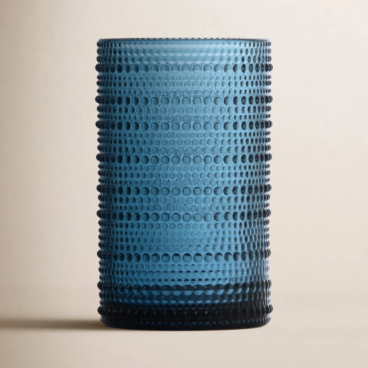 A tall, blue glass tumbler with a textured surface featuring a pattern of small circular holes, placed on a light-colored background.