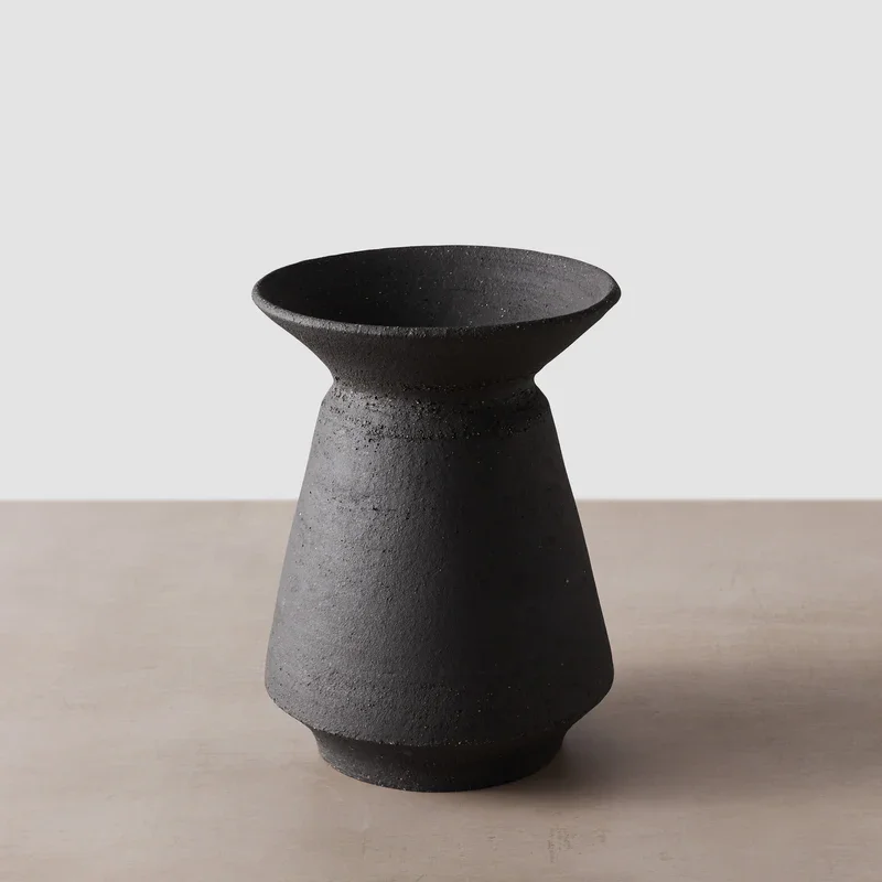 A black, textured, ceramic vase with a wide top and narrow base on a light-colored surface against a plain white background.