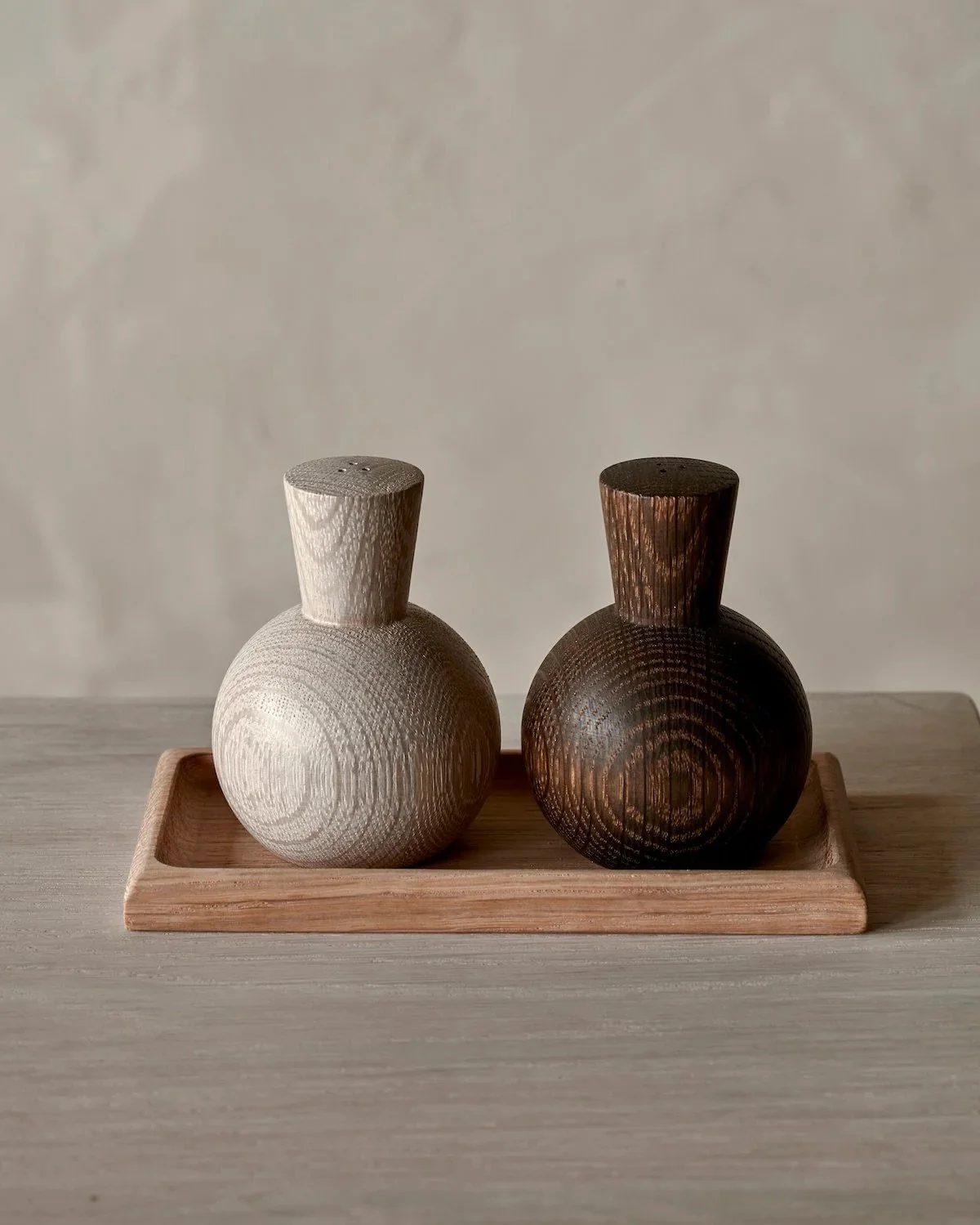 Two wooden salt and pepper shakers with rounded bases and short necks, one light-colored and one dark-colored, placed on a rectangular wooden tray on a light wooden surface.