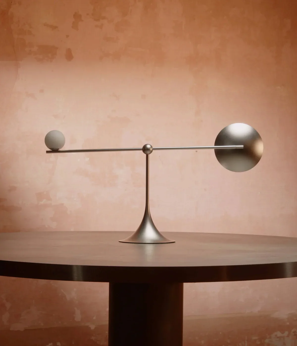 A modern, abstract cordless light with metallic spheres on a thin horizontal rod and vertical stand, mounted on a wooden table against a textured, reddish-brown wall.