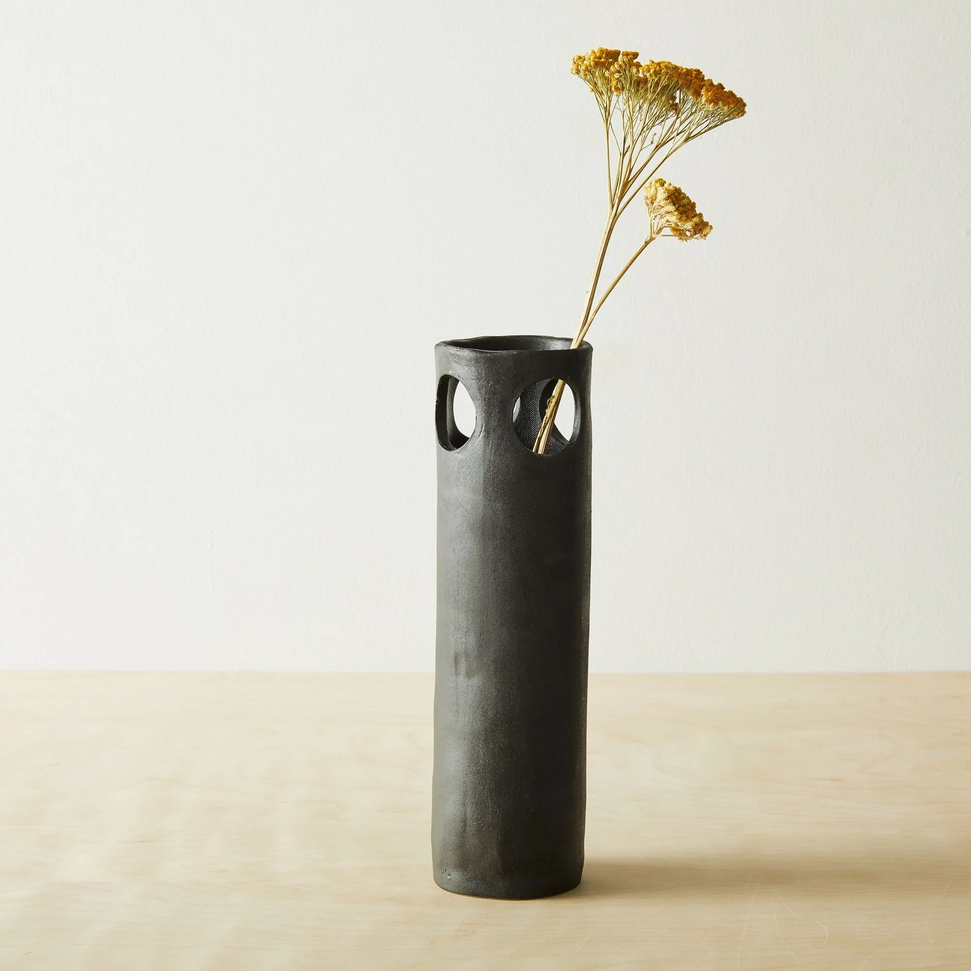 A tall black ceramic vase with round cutout handles, holding dried yellow flowers, on a light wooden surface against a plain light background.