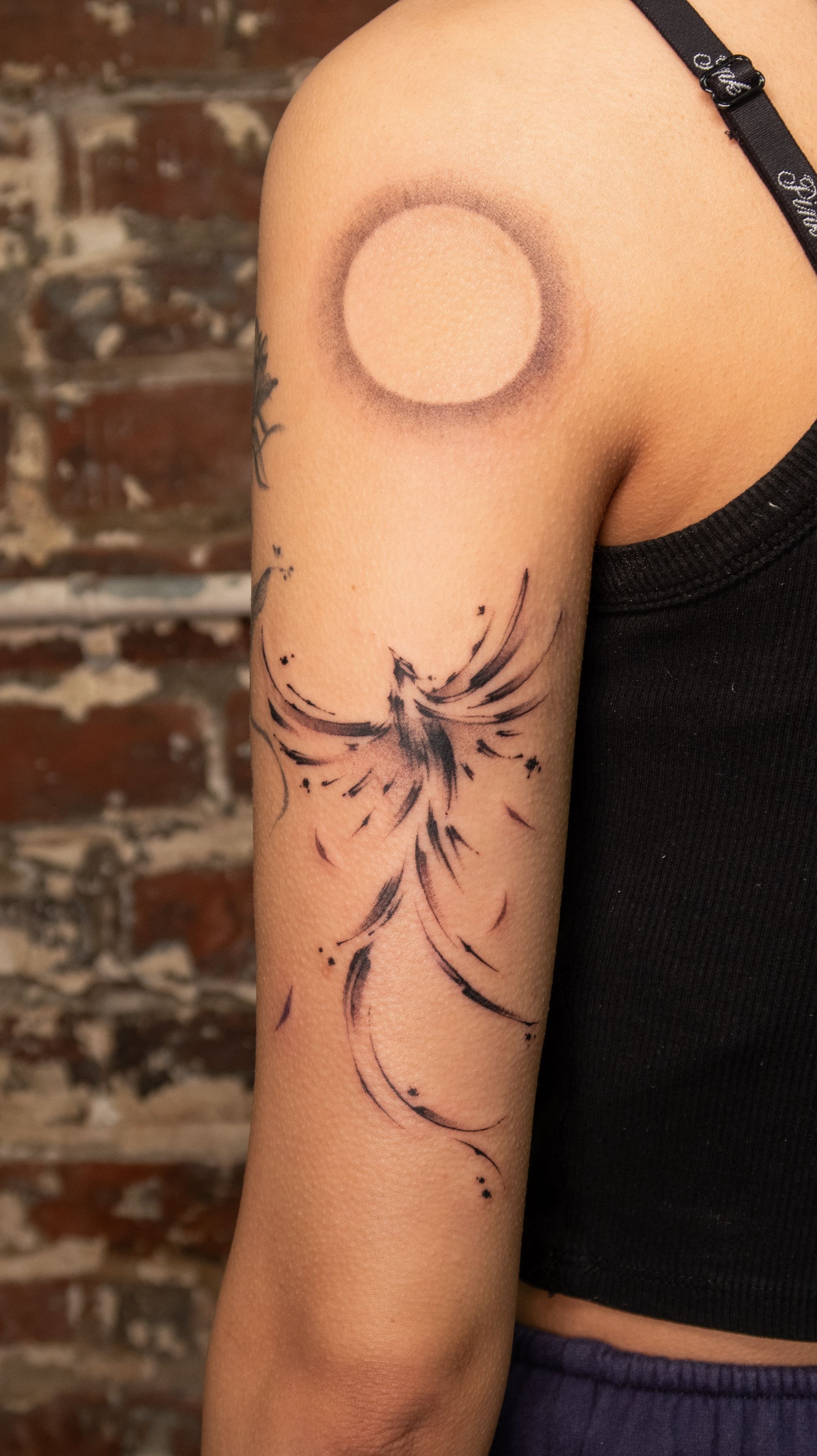 A tattoo of a stylized phoenix with black ink on a person's upper arm, with a circular tattoo surrounding the shoulder area, and a brick wall in the background.