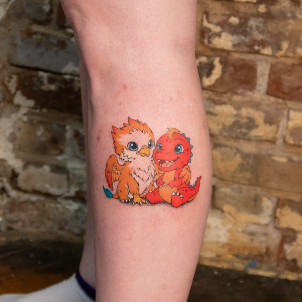 Colorful cartoon tattoo of a baby dragon and a baby bird sitting together on a person's thigh against a brick wall background.