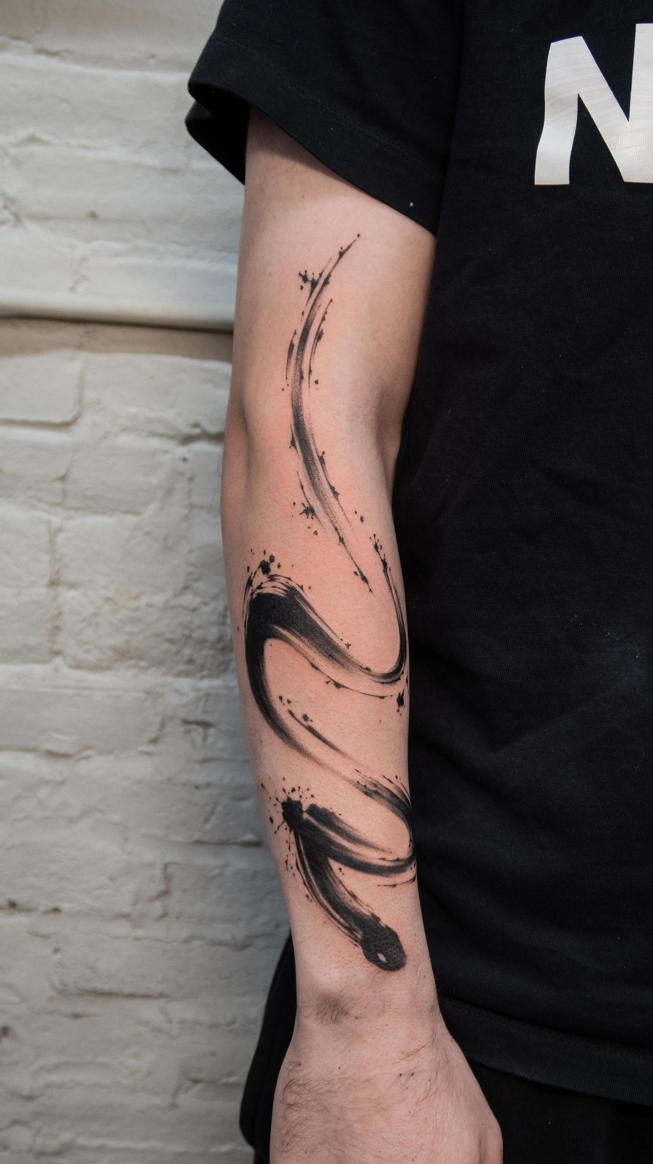 Person with a black ink abstract tattoo on their forearm, wearing a black T-shirt, standing against a white brick wall.